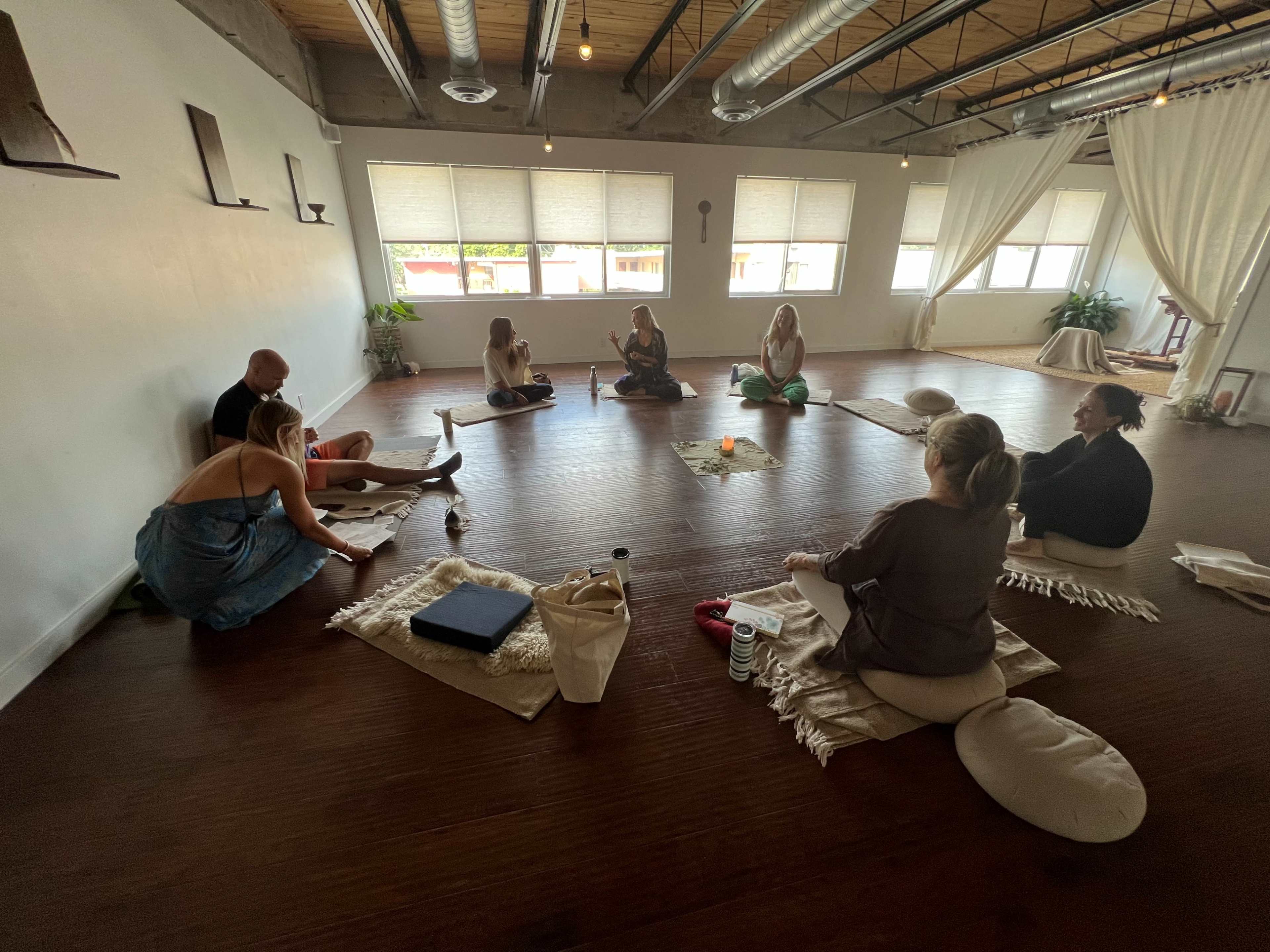 A group of individuals sits in a circle on the floor of a spacious, well-lit room, engaged in a meditation or reflective session.