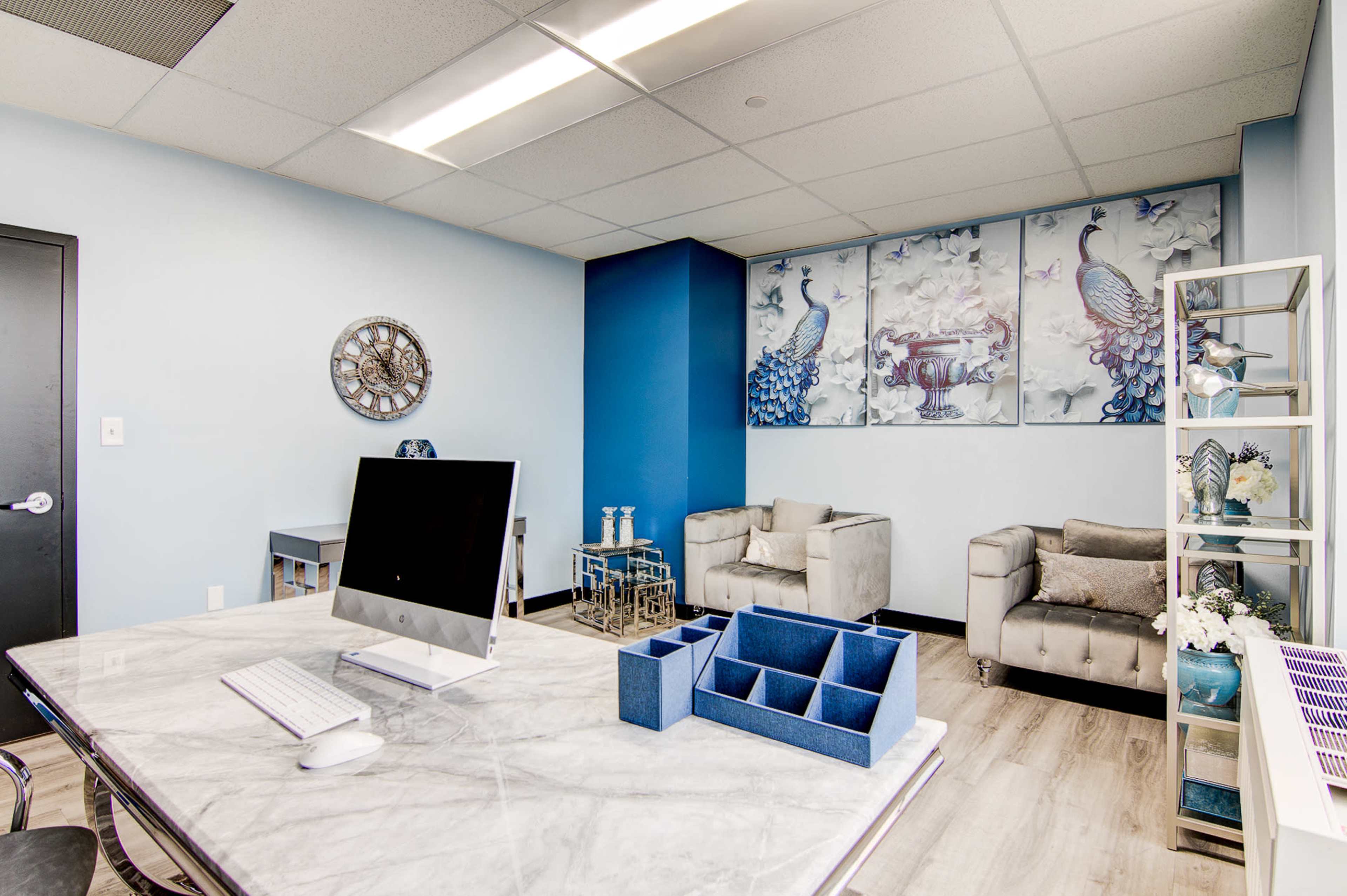 A modern office features a marble desk with a computer, blue accents, and two grey sofas against a wall adorned with peacock artwork.