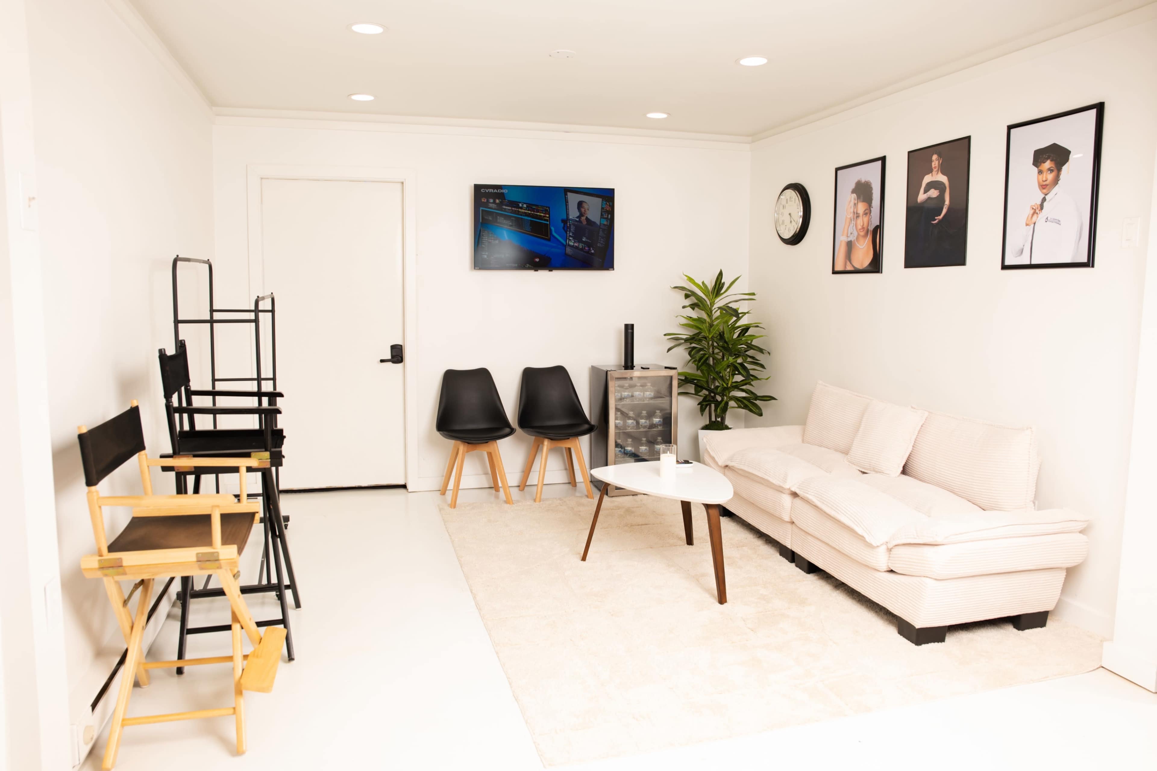 The image shows a modern, minimalist lounge area featuring a beige sofa, two black chairs, a small coffee table, and various framed photographs on the wall.