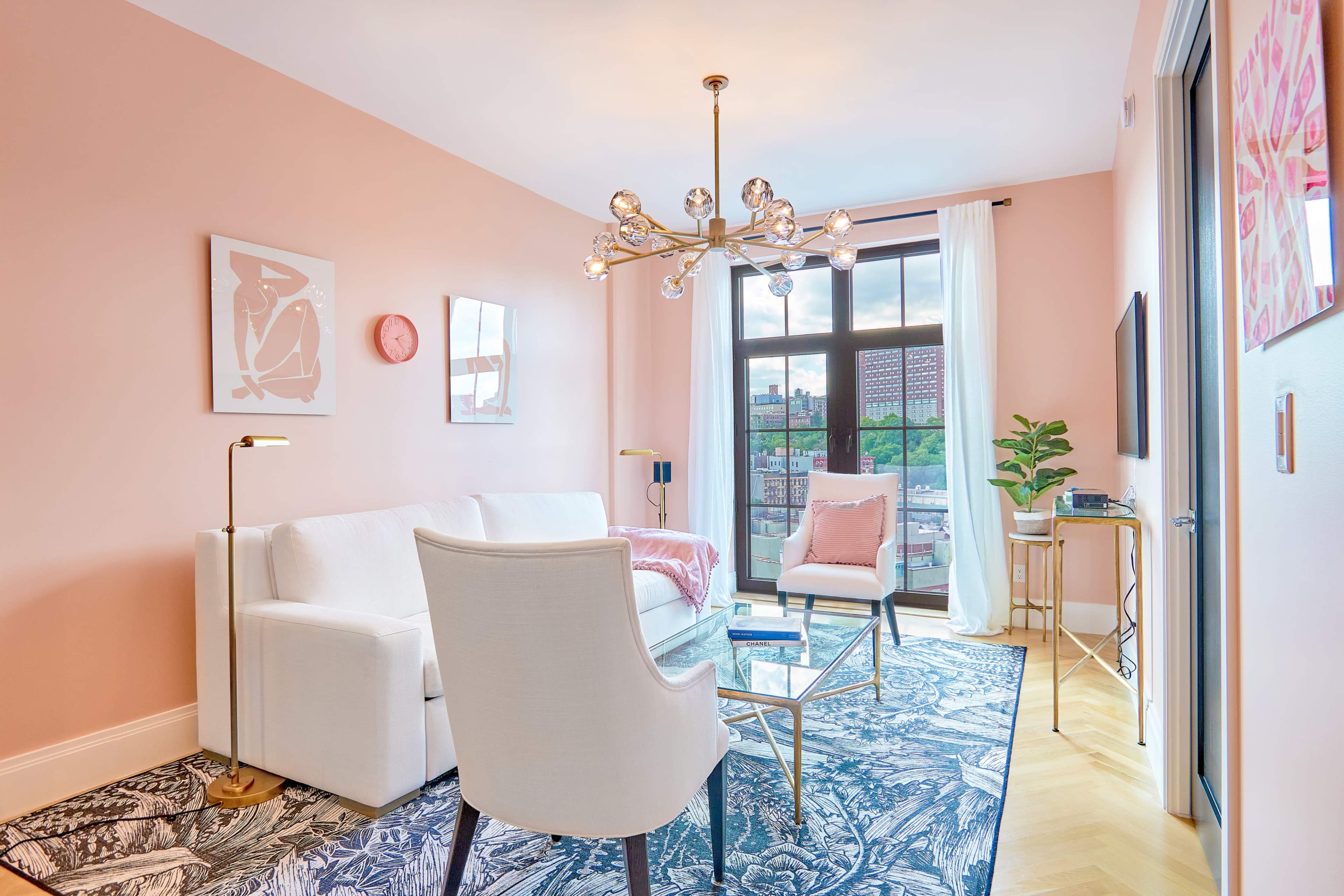A modern living room features a white sofa, a glass coffee table, and large windows with sheer curtains, surrounded by light pink walls and decorative artwork.