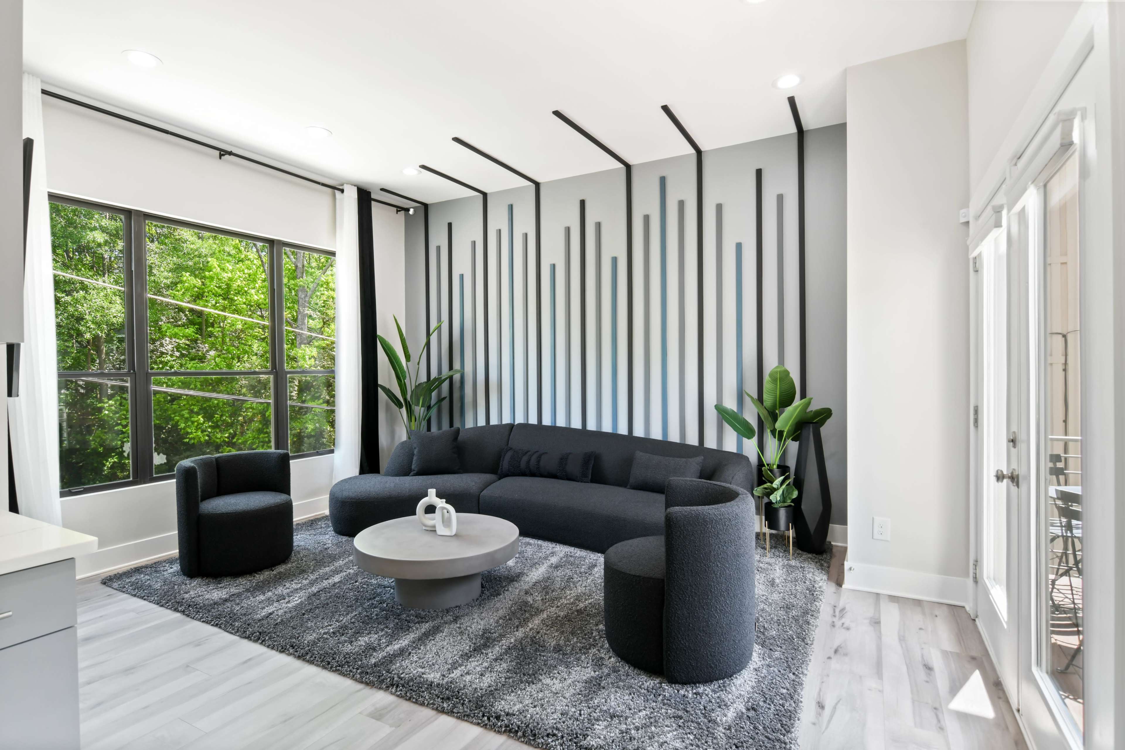 A modern living room features a black sectional sofa, round coffee table, and a textured gray rug, with large windows letting in natural light.