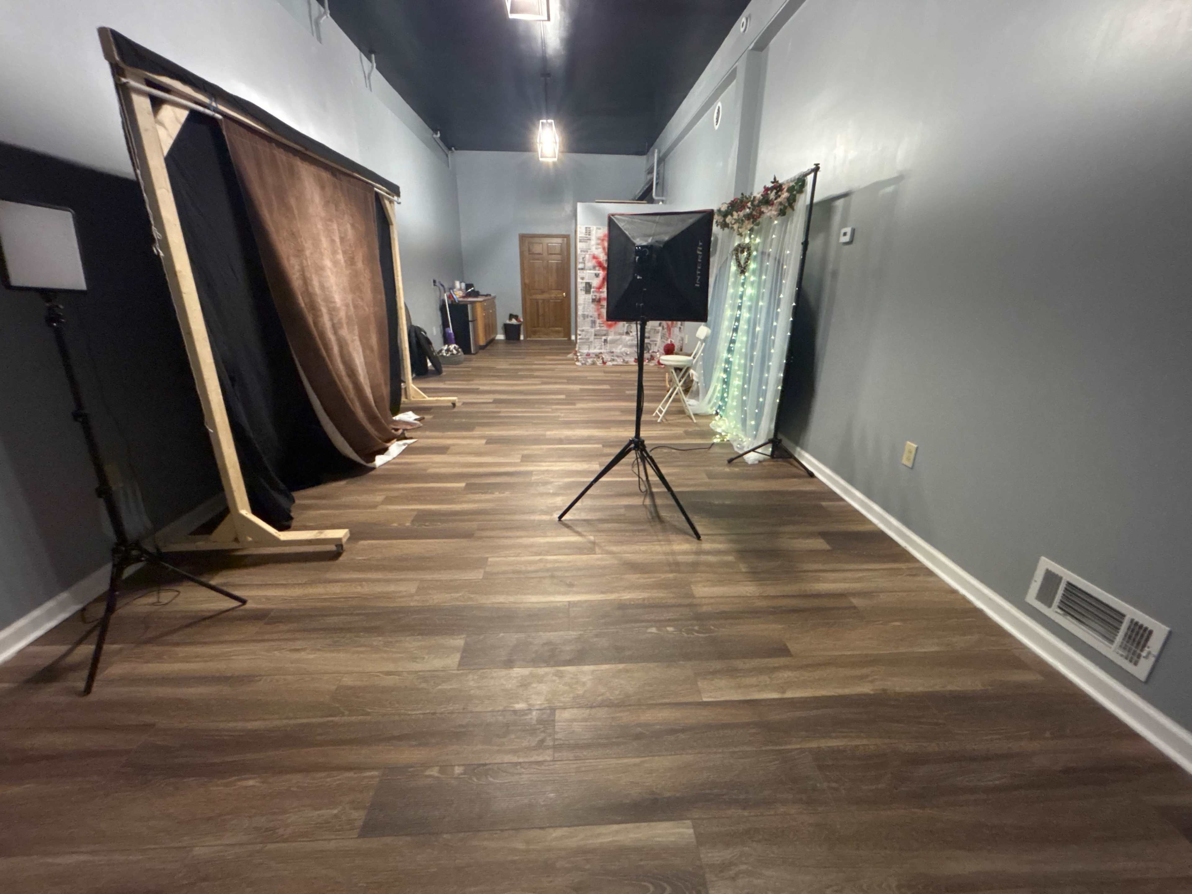 The image shows an empty interior space with wooden flooring, featuring a backdrop setup, light stands, and decorative elements along one side.