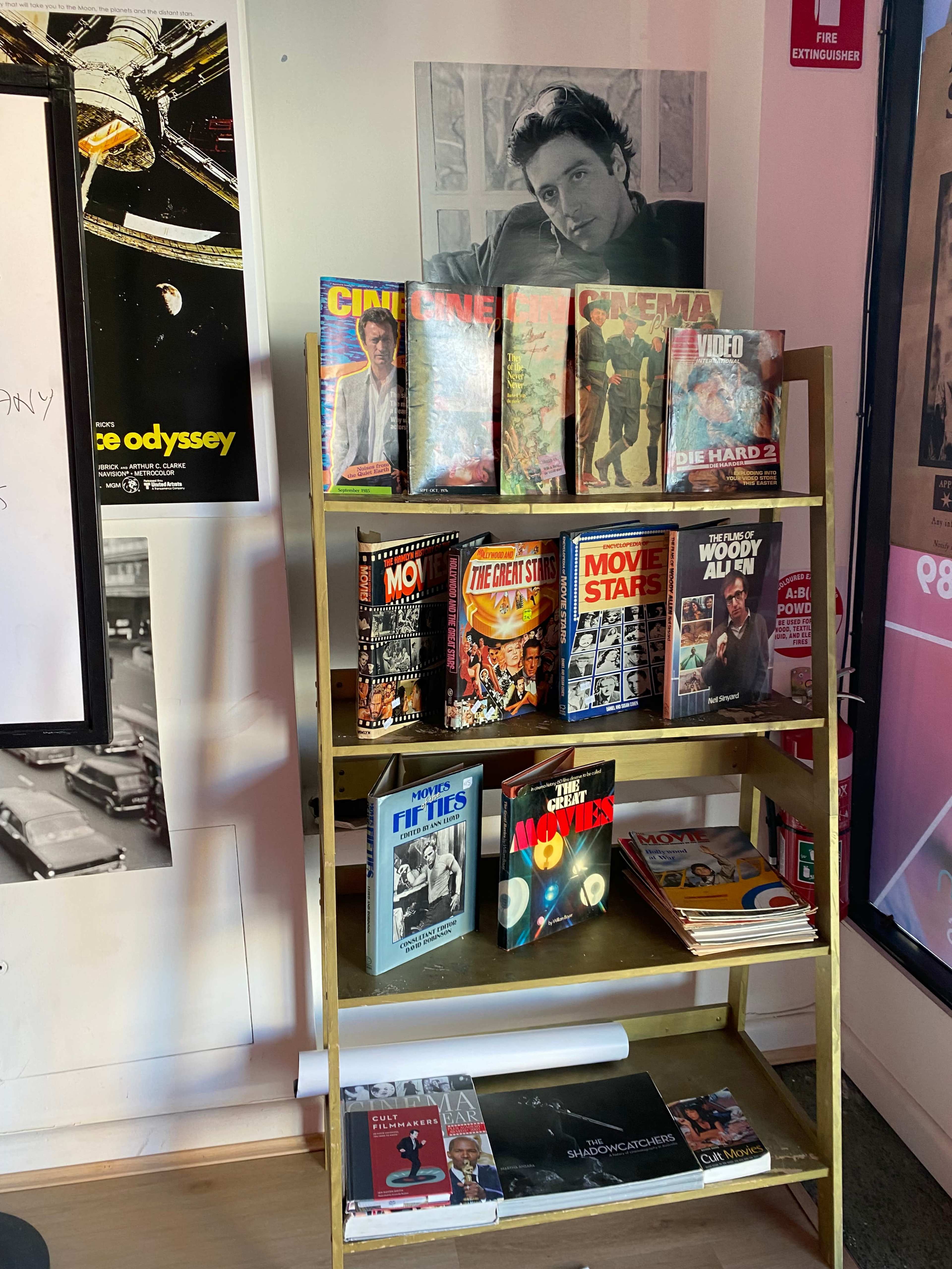 The image shows a gold metal bookshelf holding several magazines related to cinema, stacked against a backdrop featuring a poster of a film and some film-themed decor.