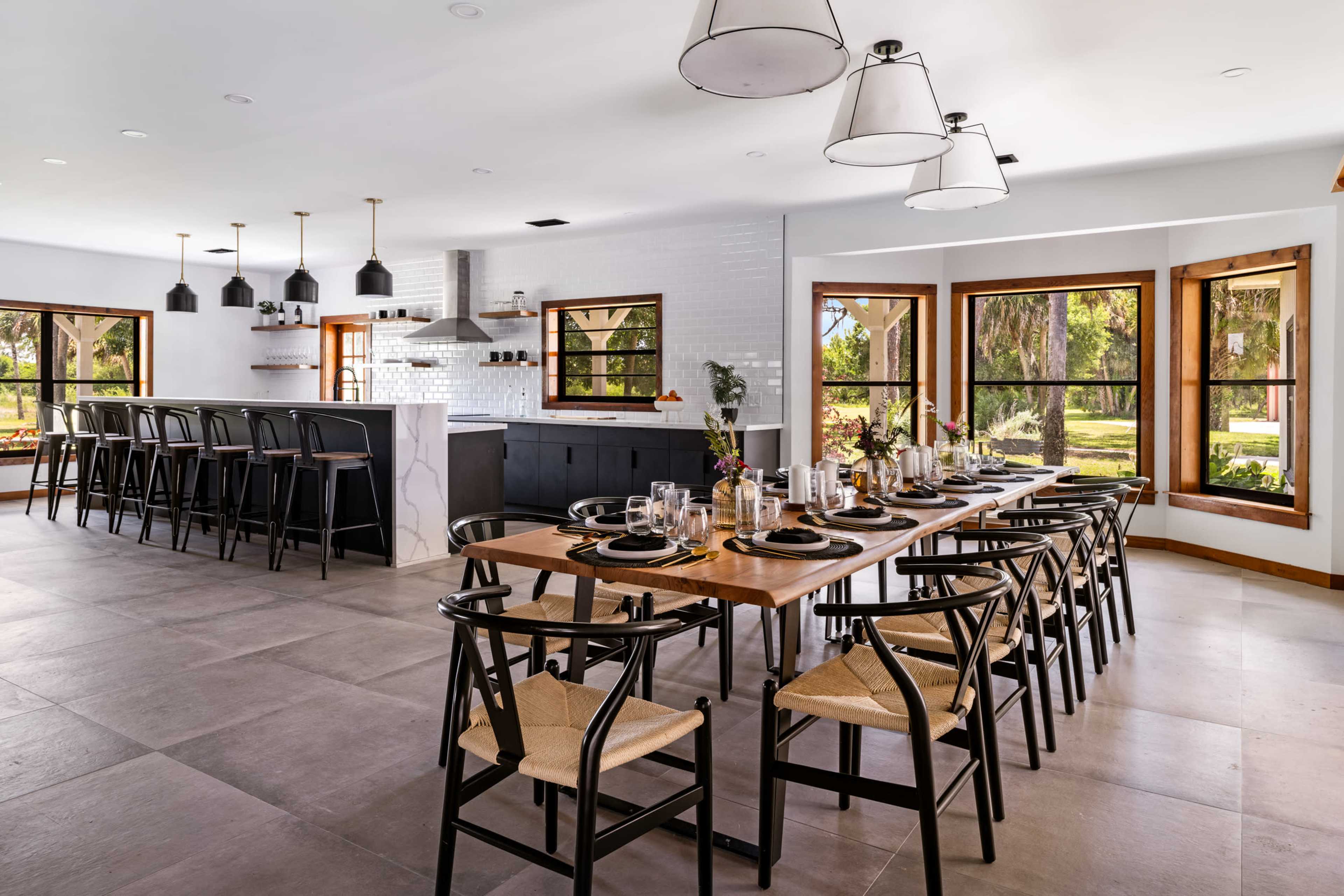 A modern kitchen and dining area with a large wooden table set for a meal, surrounded by black chairs, and featuring large windows that allow natural light to fill the space.