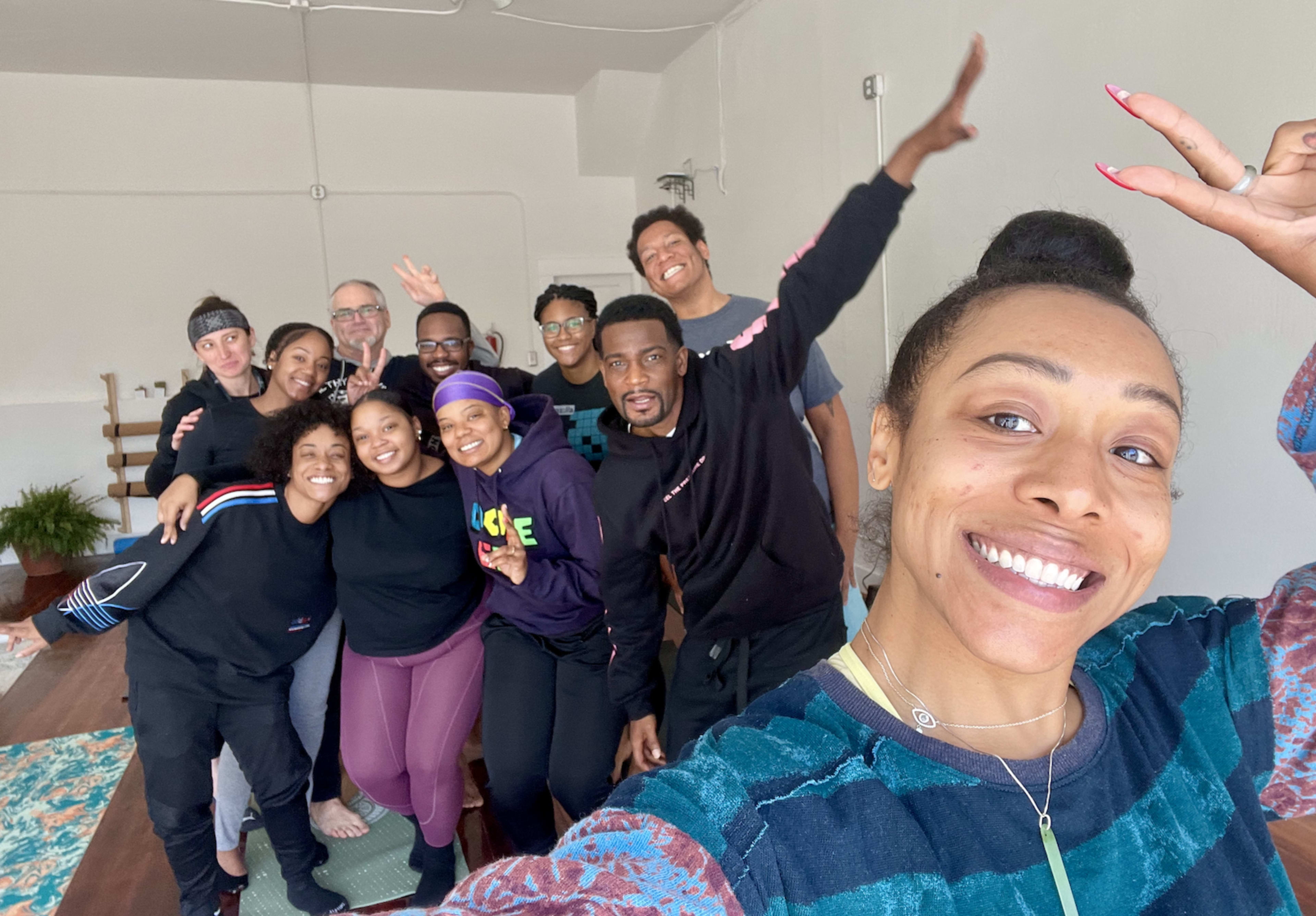 A group of twelve people smiles and poses for a selfie in a bright, spacious yoga studio.