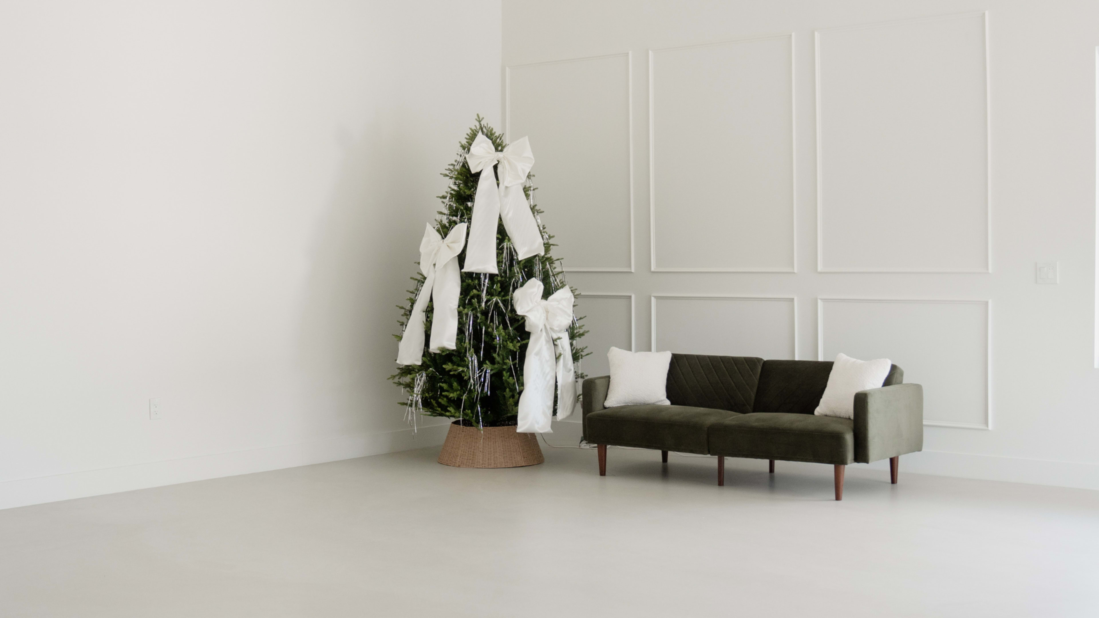 A decorated Christmas tree is placed next to a modern sofa in a minimalist room with white walls.
