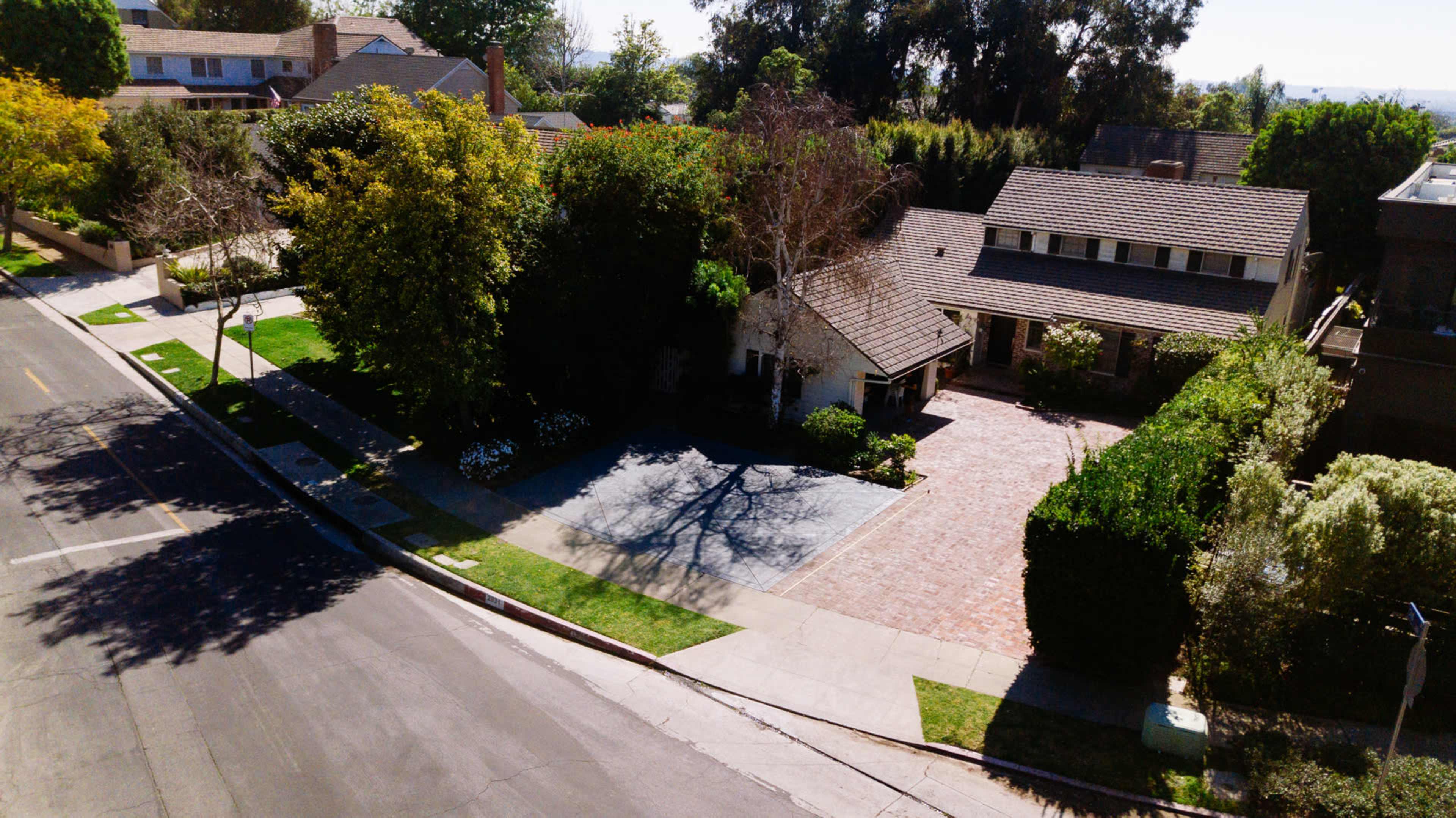 The image shows a residential neighborhood with a large house featuring a brick driveway, surrounded by well-kept lawns and trees.