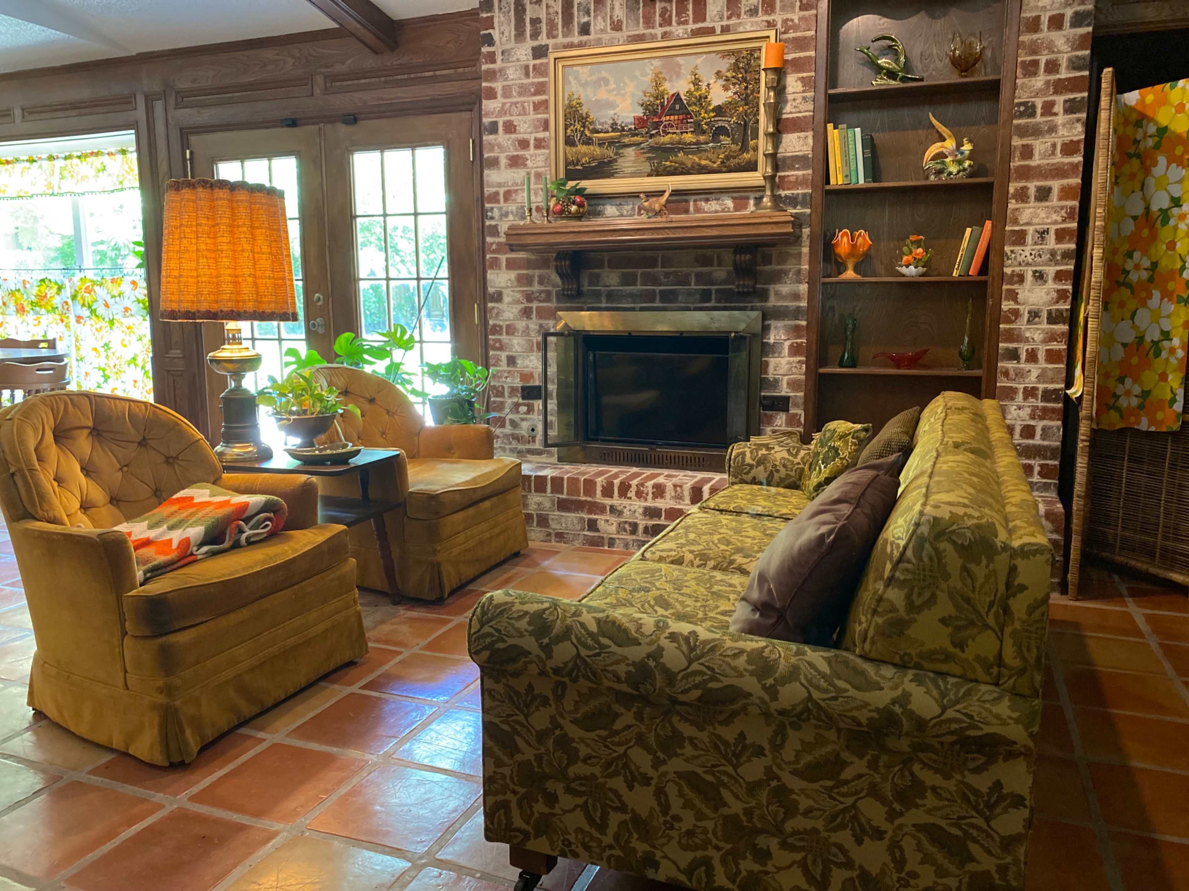 A living room features a sofa and two armchairs arranged around a coffee table in front of a brick fireplace, with a view of a colorful window and a bookshelf.
