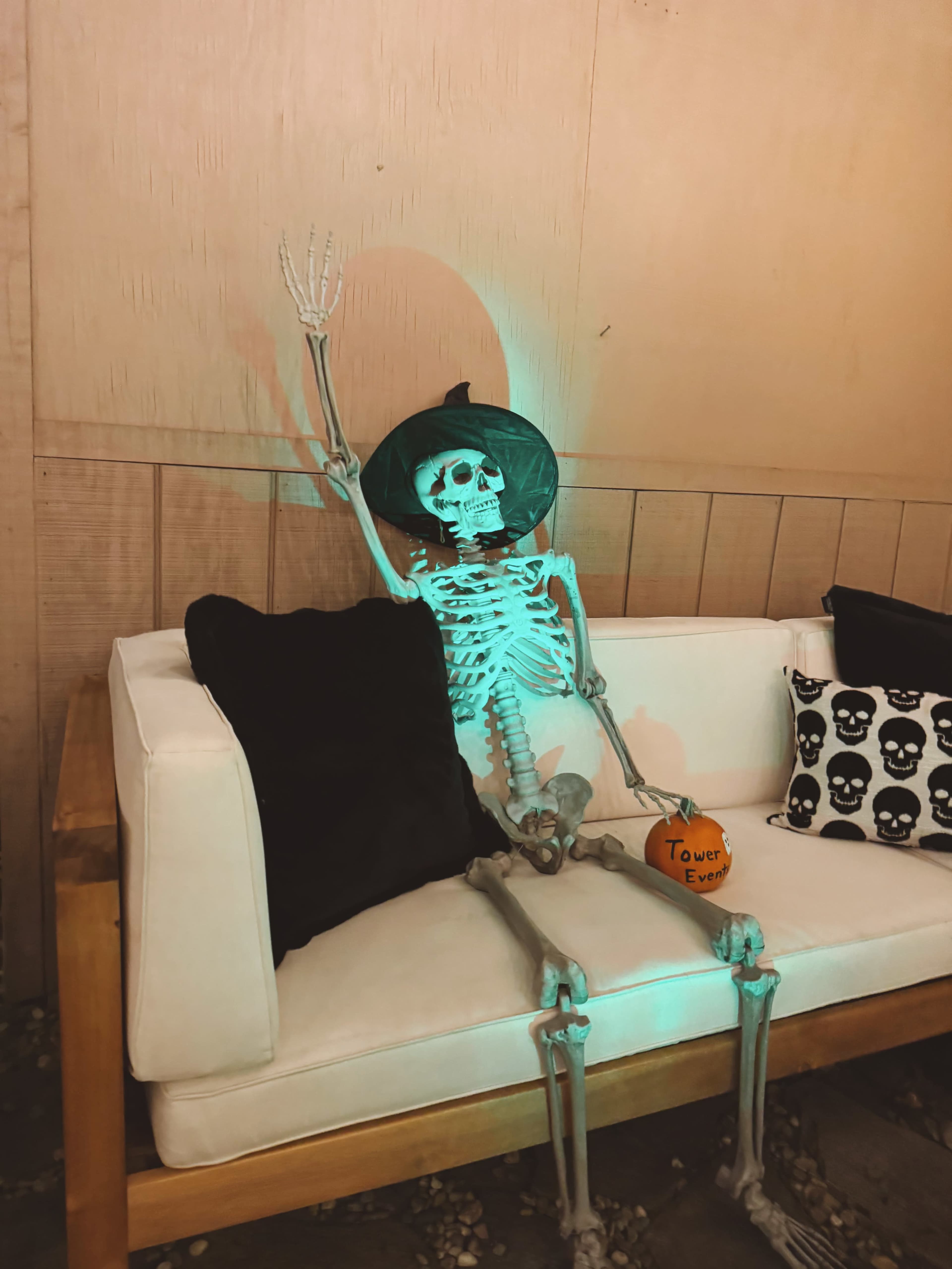A skeleton wearing a witch's hat sits on a couch with decorative pillows featuring skulls.