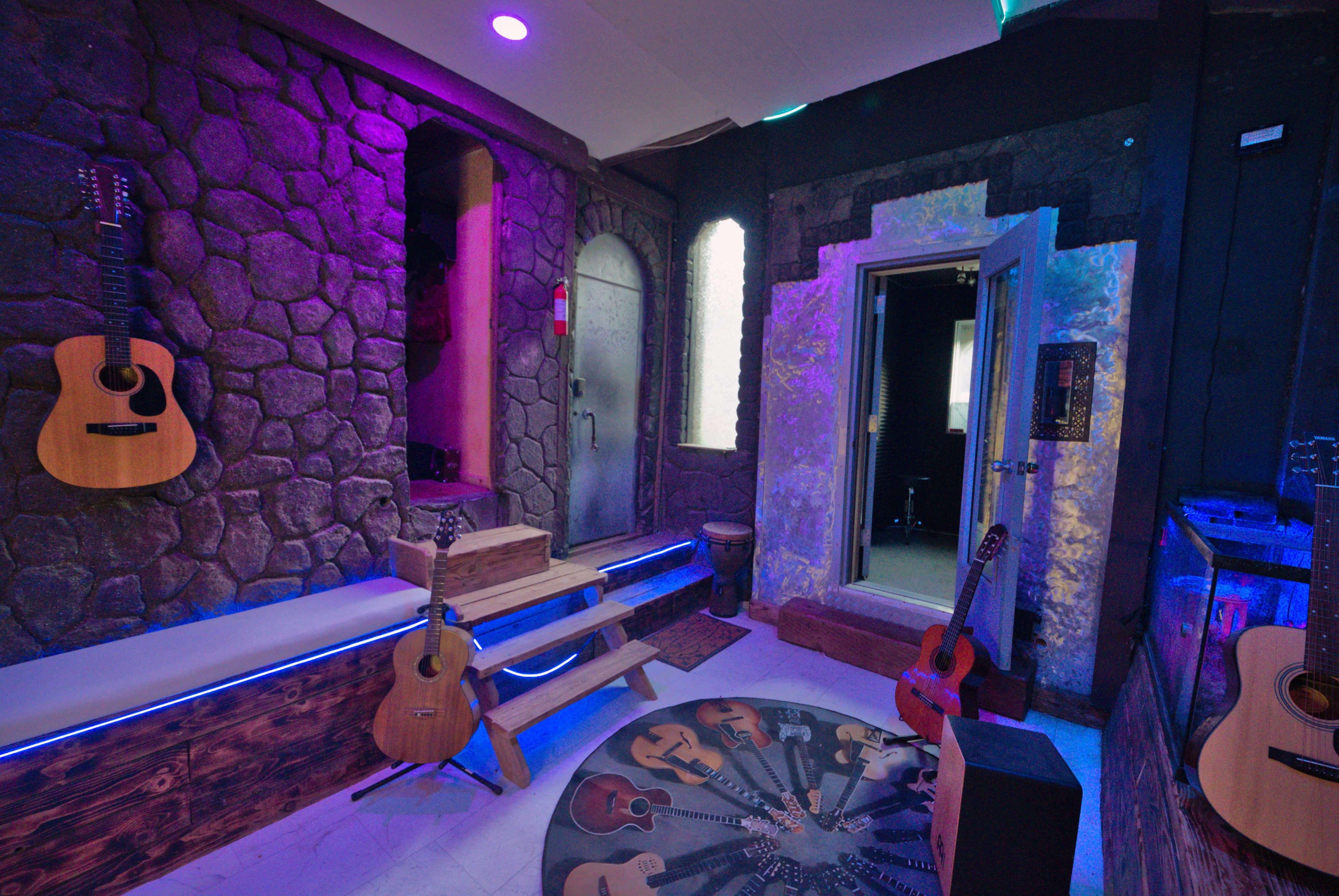 The image shows a music studio with stone walls, wooden steps, and several guitars displayed around a circular rug featuring guitar designs.