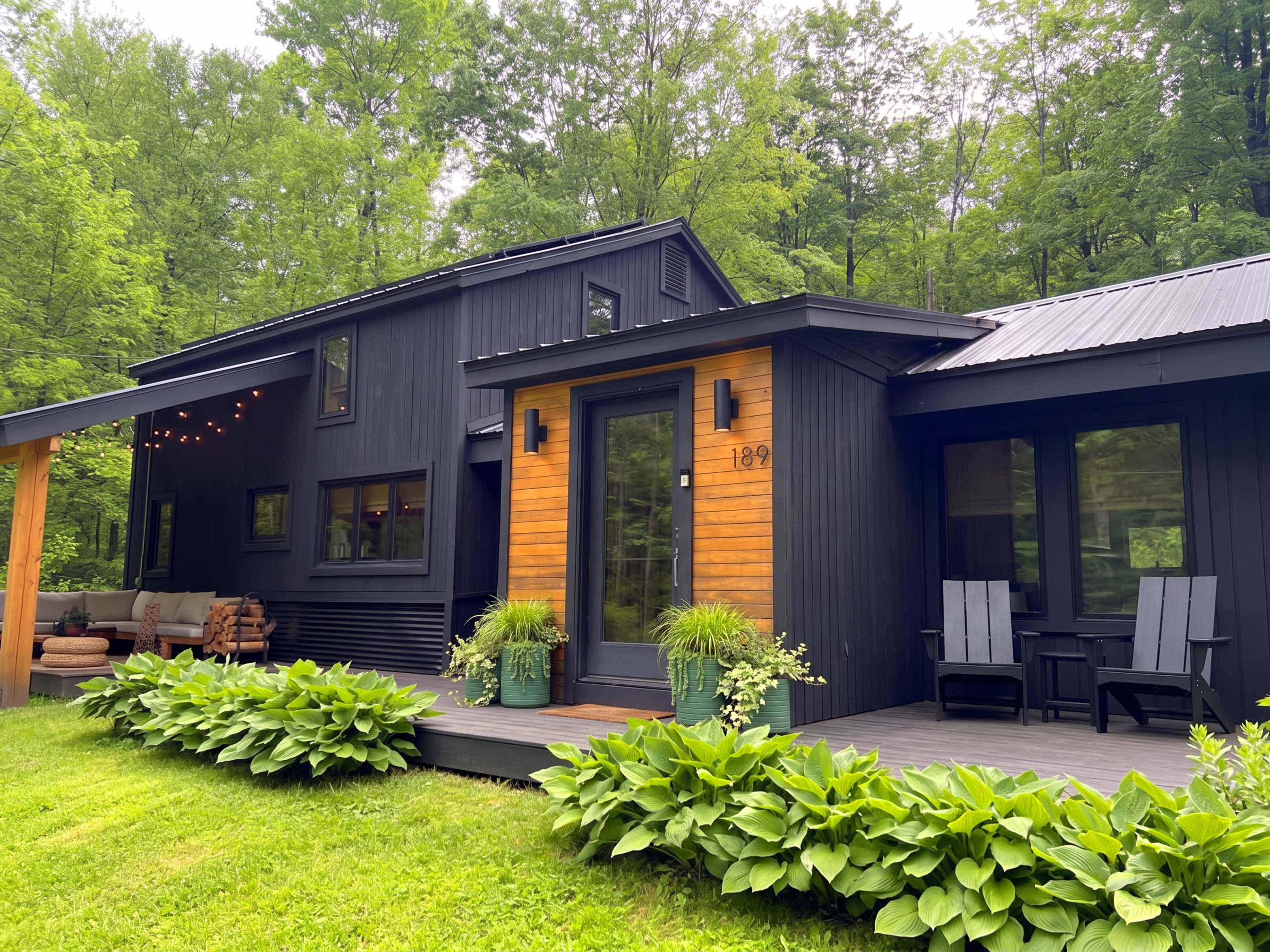 The image shows a modern black and wooden house surrounded by greenery, featuring a welcoming porch and seating area.