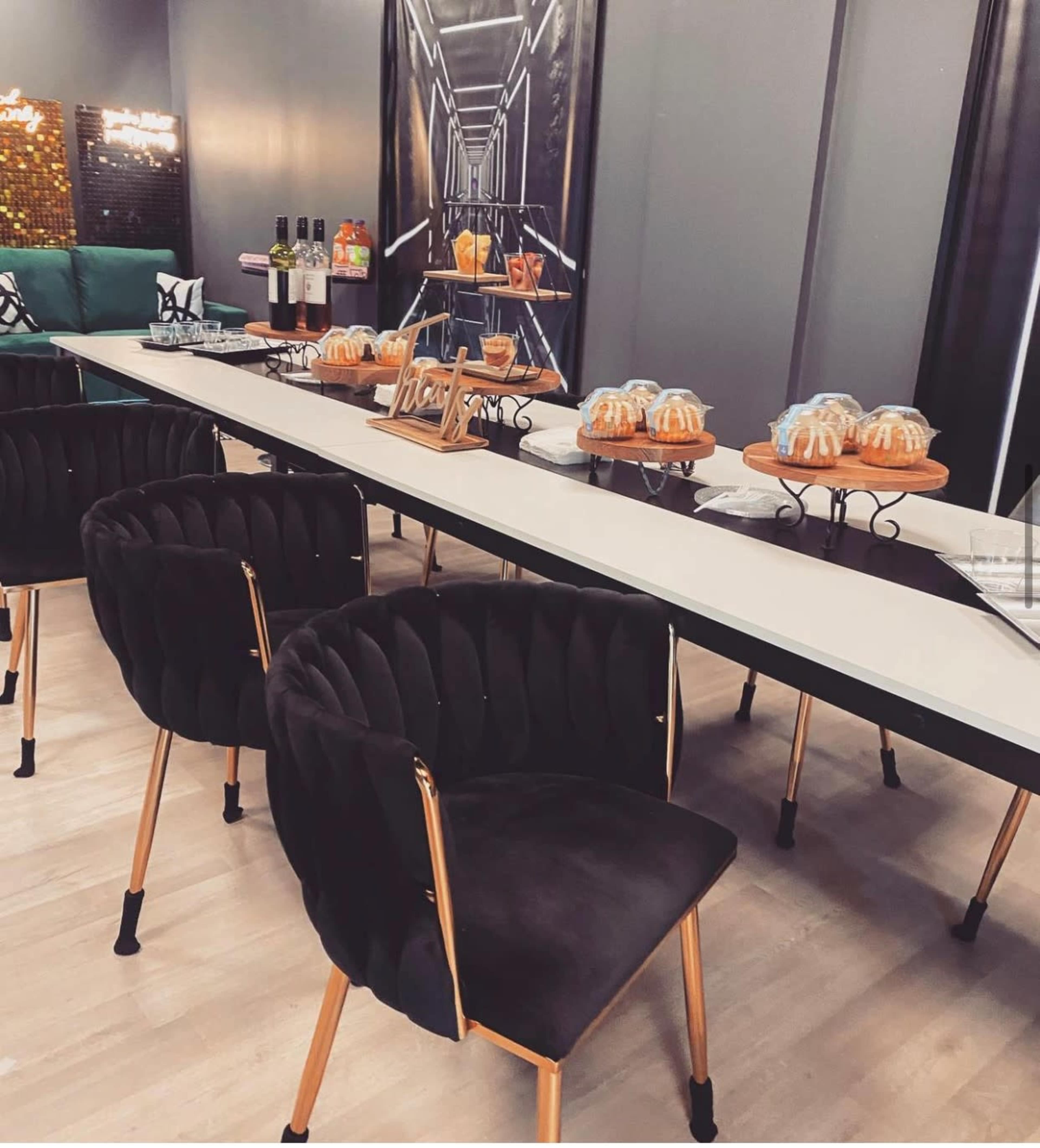 A long, modern dining table is set with decorative bundt cakes and surrounded by velvet chairs with gold legs in a stylish interior setting.