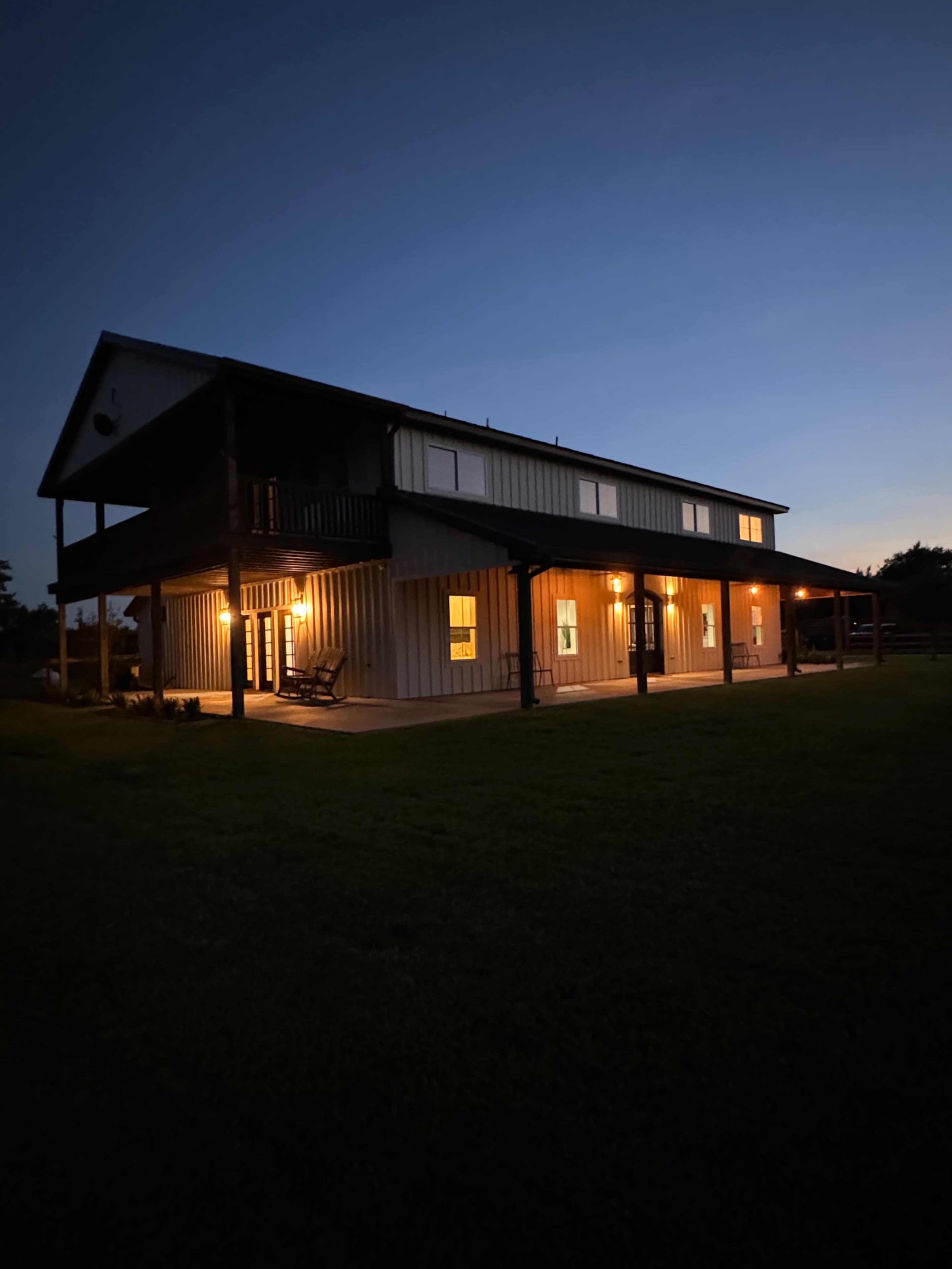 A two-story house with exterior lighting and large windows stands on a grassy lawn during twilight.
