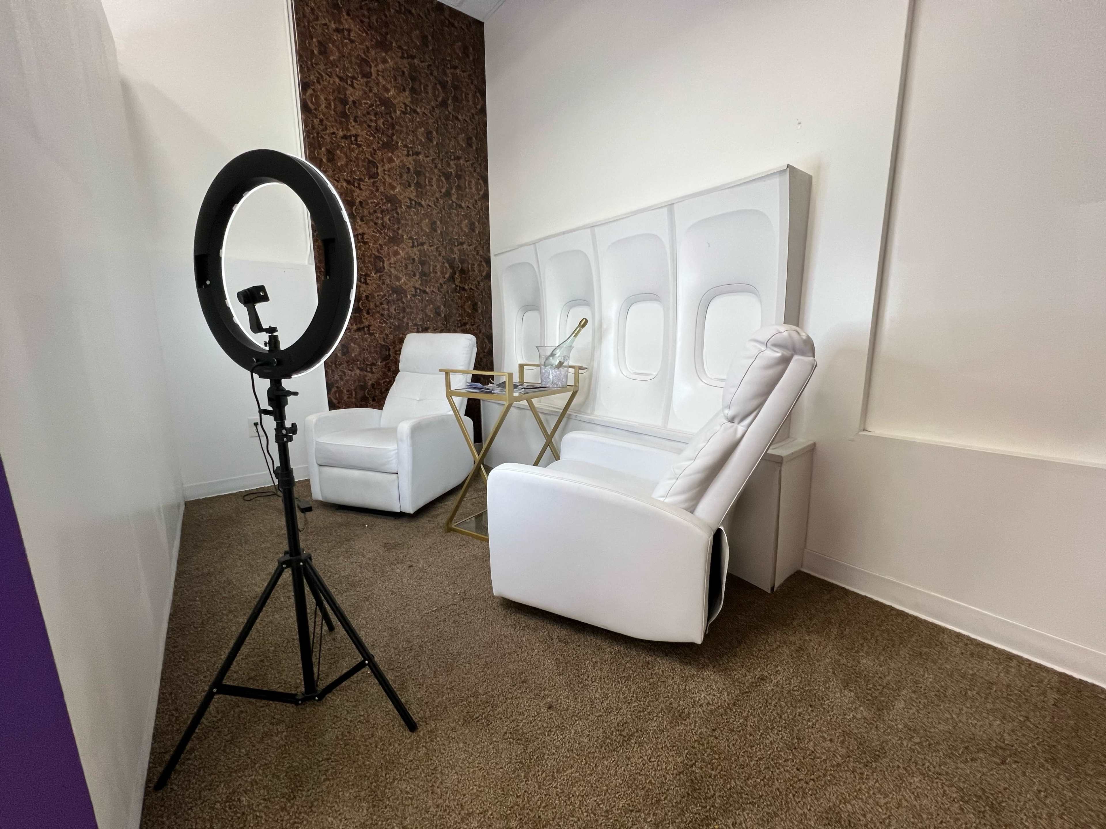 A small room with two white reclining chairs, a gold side table, and a ring light set up on a tripod.