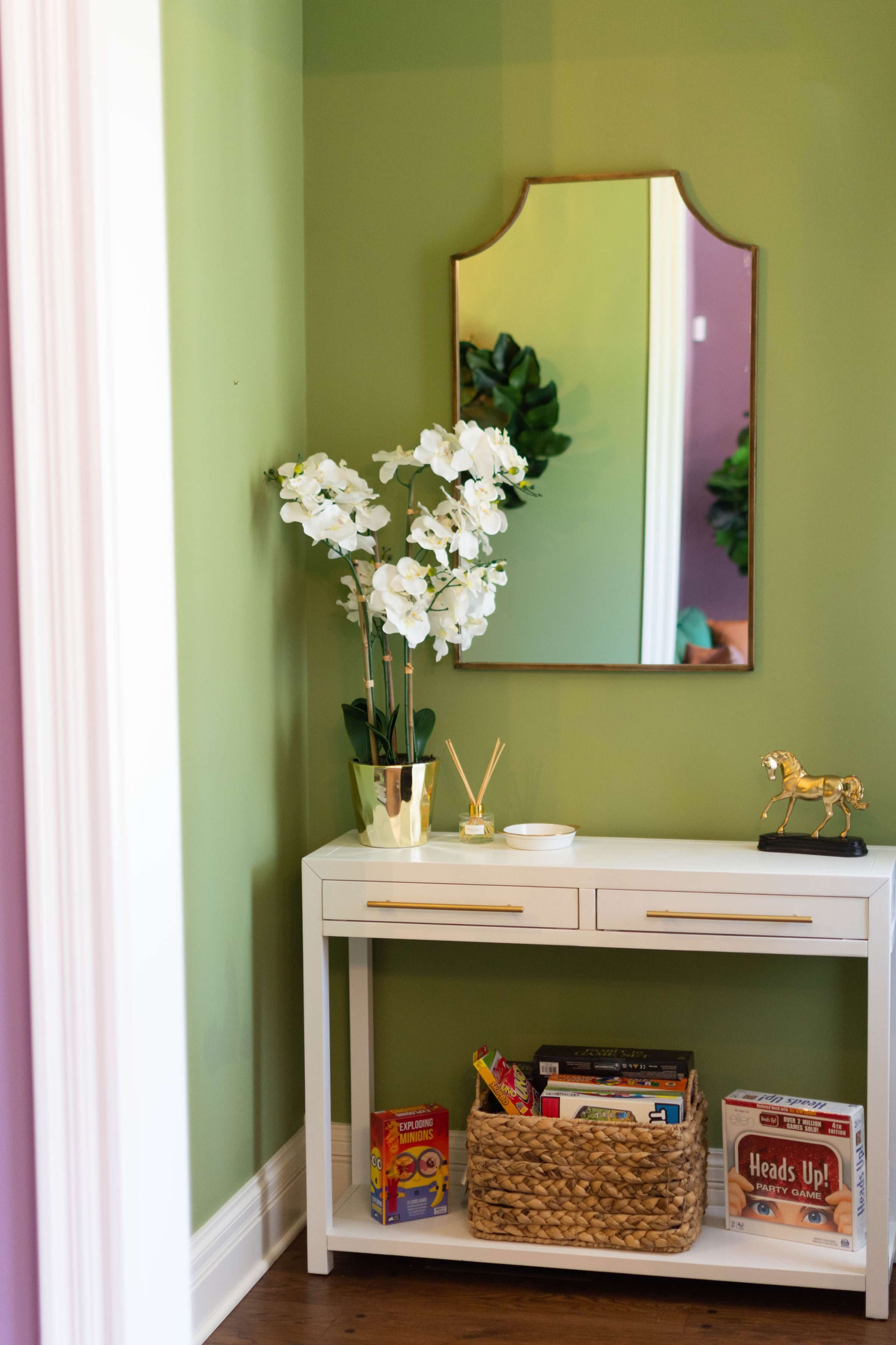 A white console table with drawers, topped by a mirror and decorative items, is positioned against a green wall adorned with potted plants and flowers.