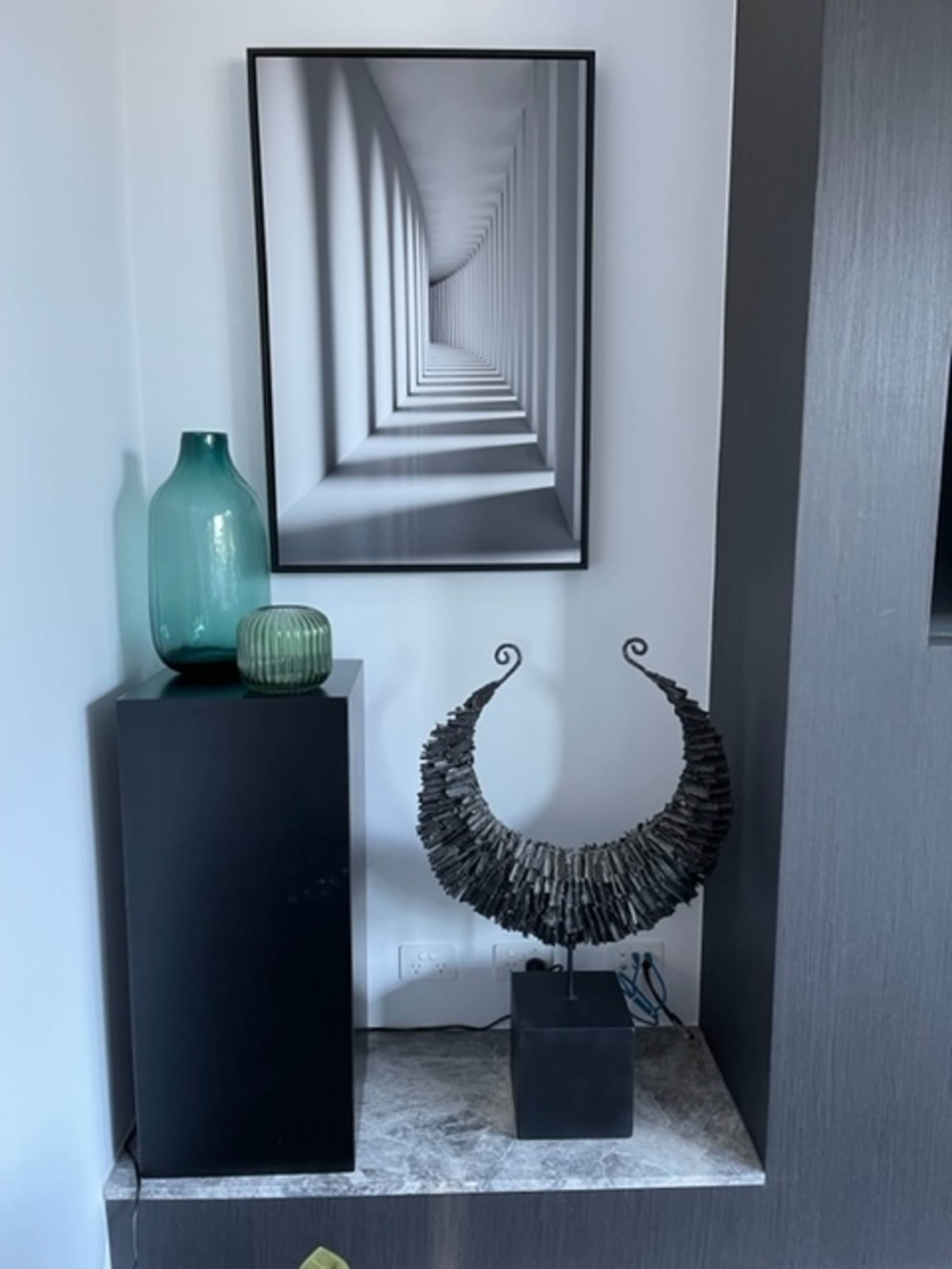 A corner of a room featuring a large framed black-and-white photograph, a green glass vase, and a decorative sculpture on a pedestal.