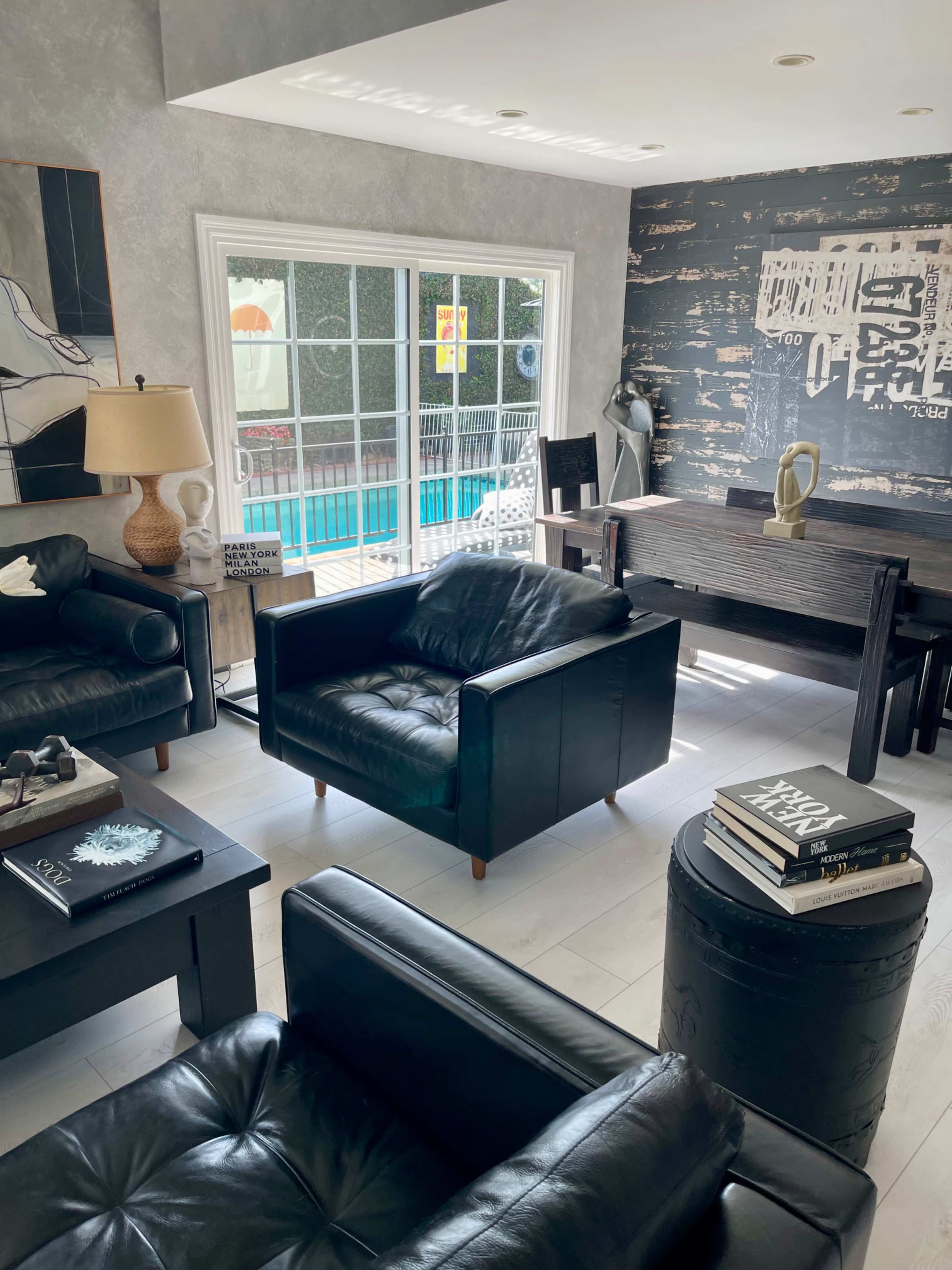 The image shows a modern living room featuring black leather furniture, a wooden table, and a view of a swimming pool through large windows.