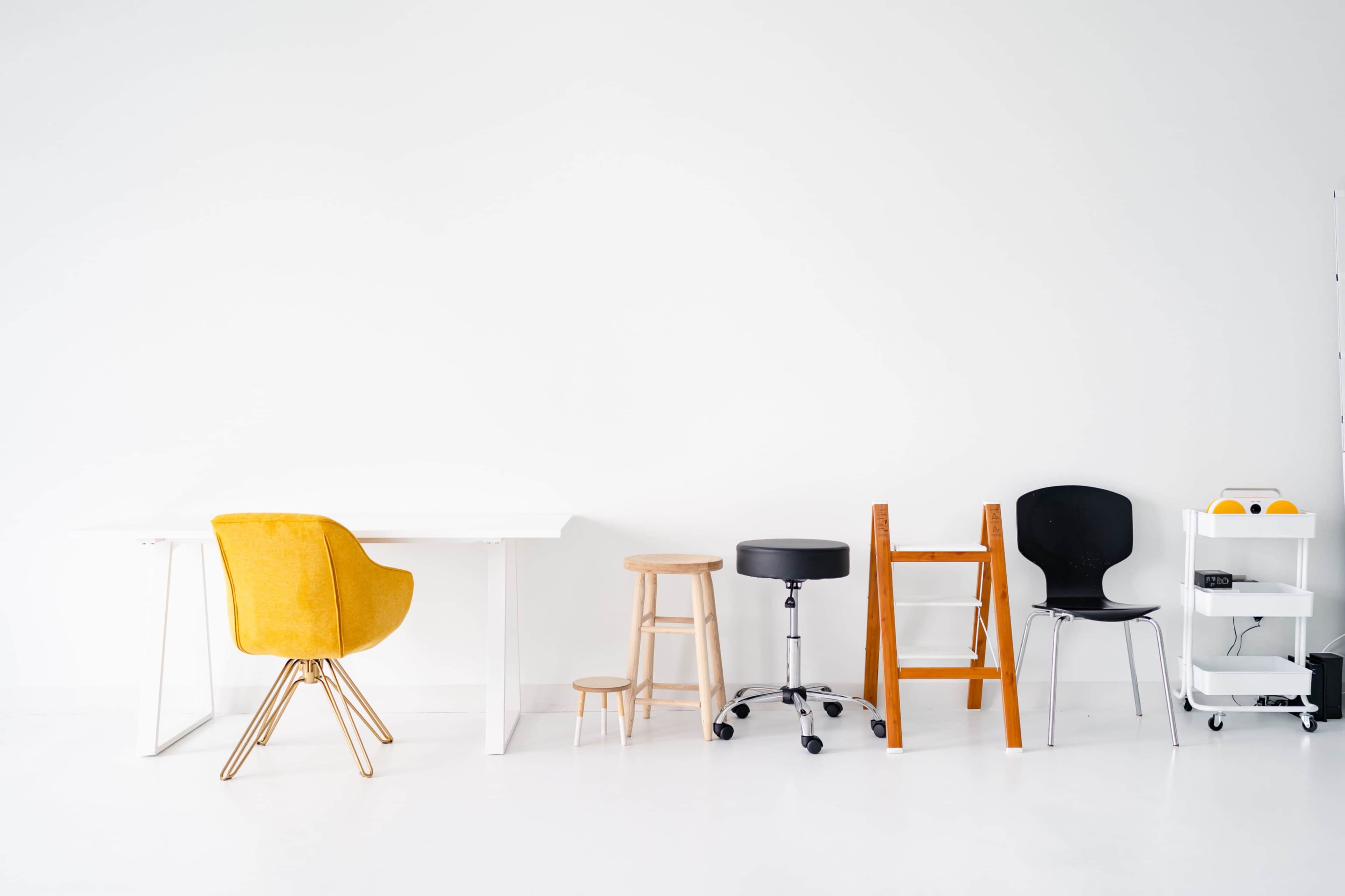 A variety of chairs and stools are lined up against a white wall, alongside a simple white table.