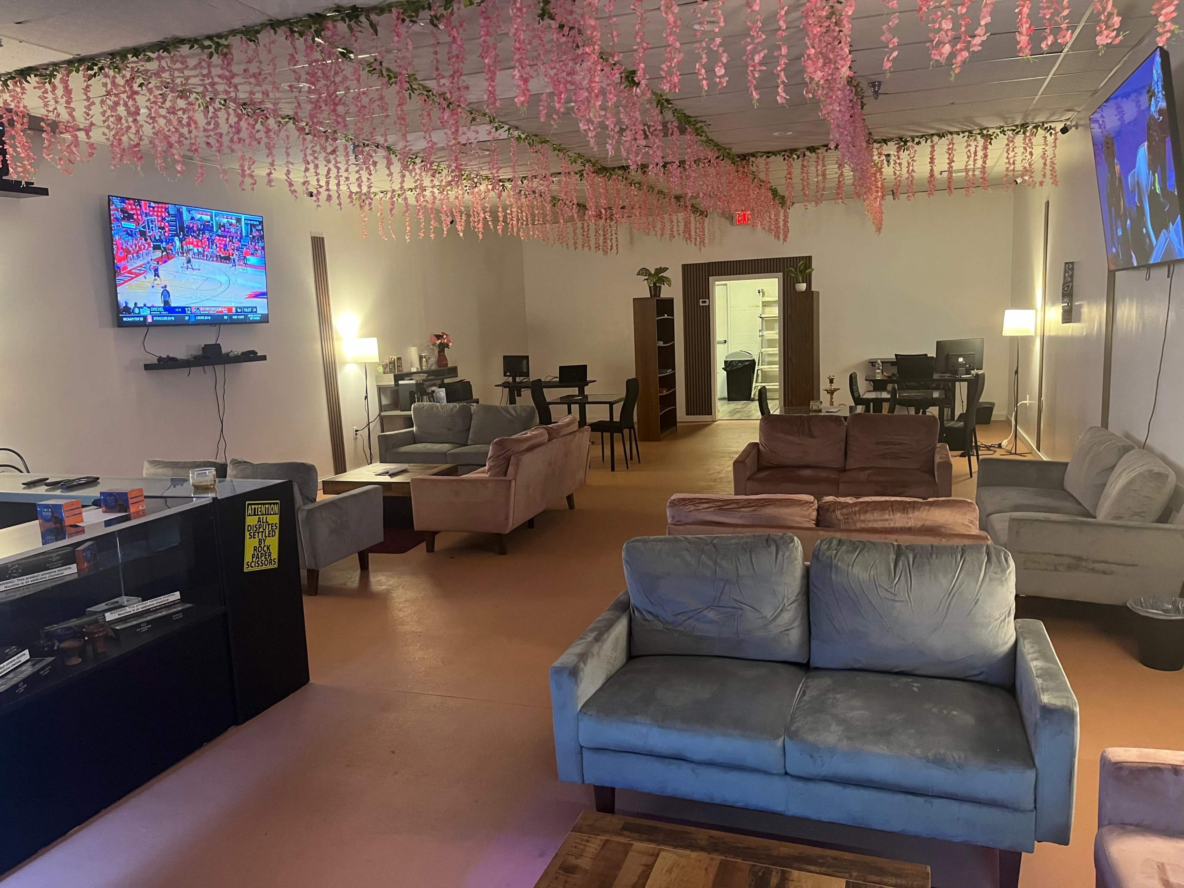 The image shows a spacious lounge area with several sofas, a television displaying a basketball game, and workstations along the walls, all decorated with hanging floral arrangements.