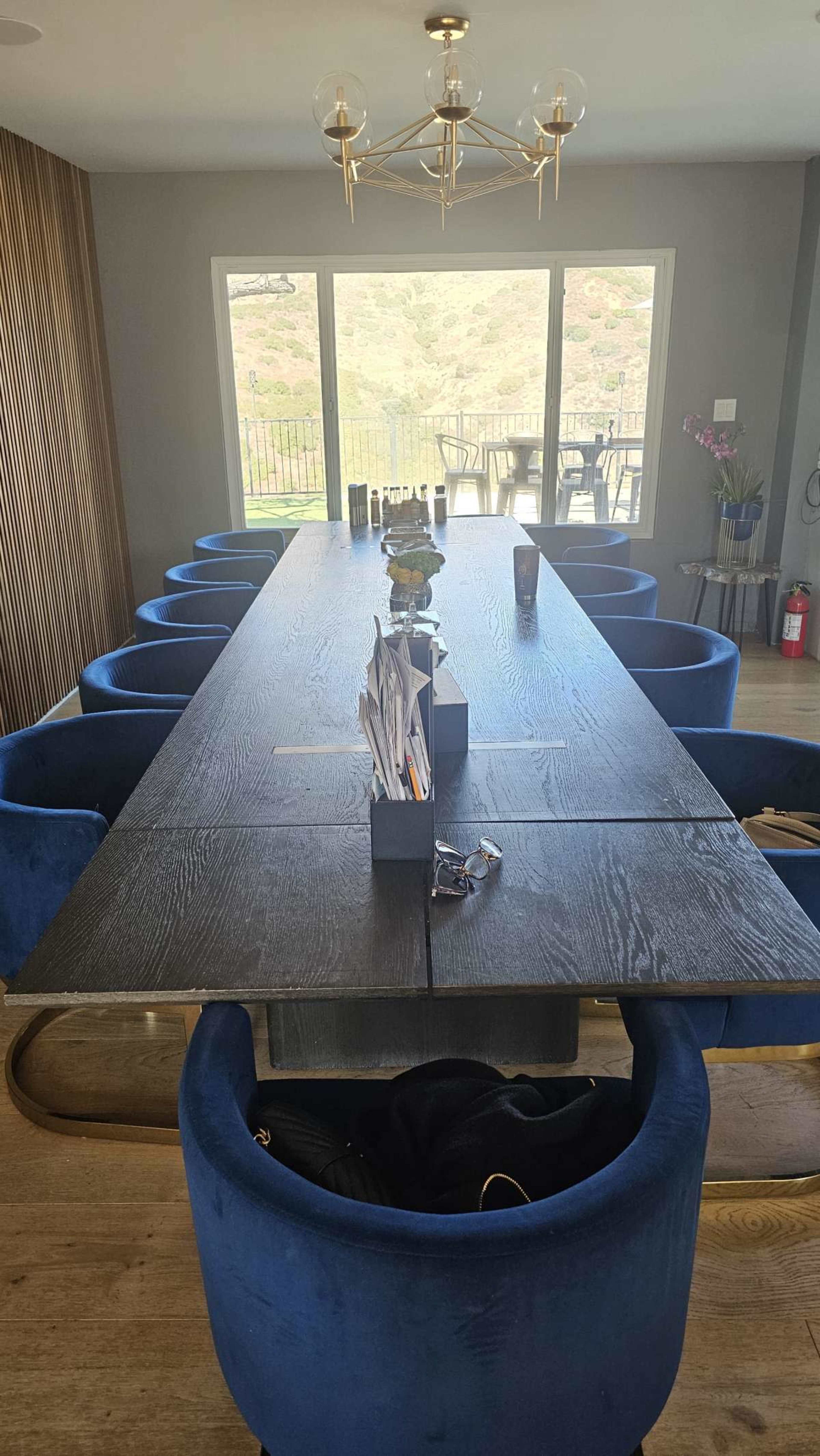 A long wooden dining table with eight blue upholstered chairs is set in a bright room with large windows overlooking a green landscape.