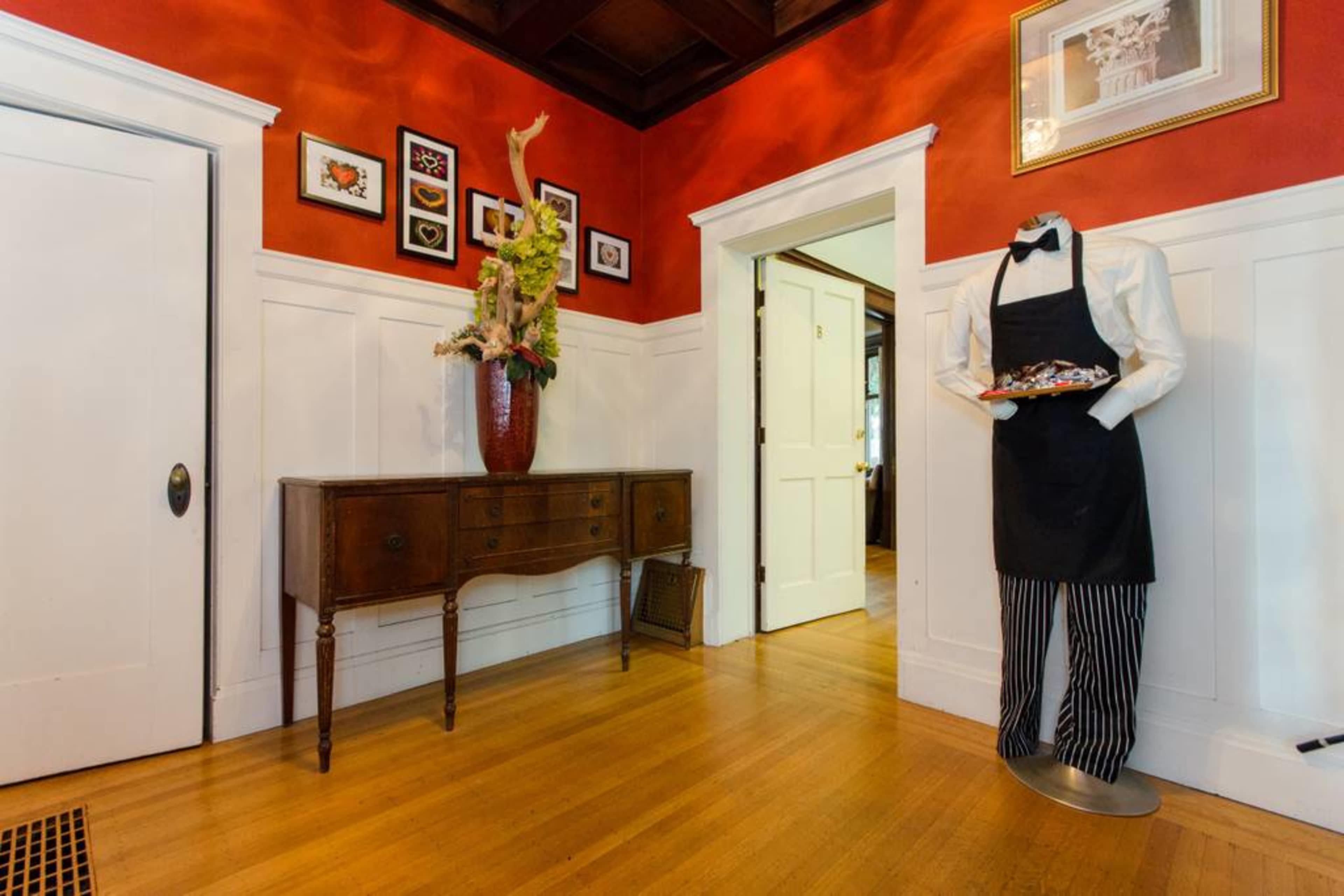 The image shows a foyer with a red wall, a wooden side table, a vase of flowers, and a mannequin dressed in formal attire holding a tray.