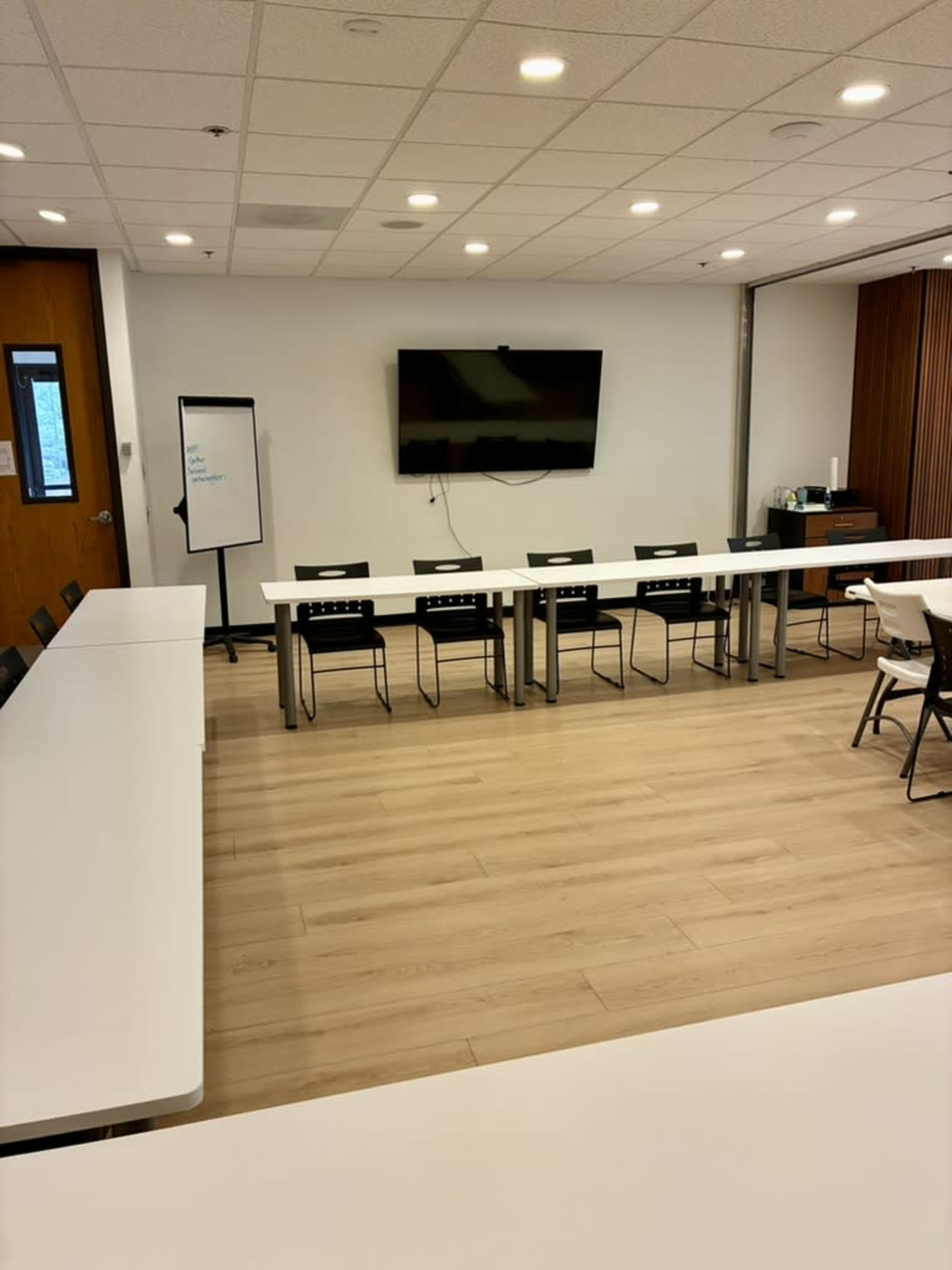 The image shows a spacious meeting room with a long white table, several chairs, a television mounted on the wall, and a whiteboard.