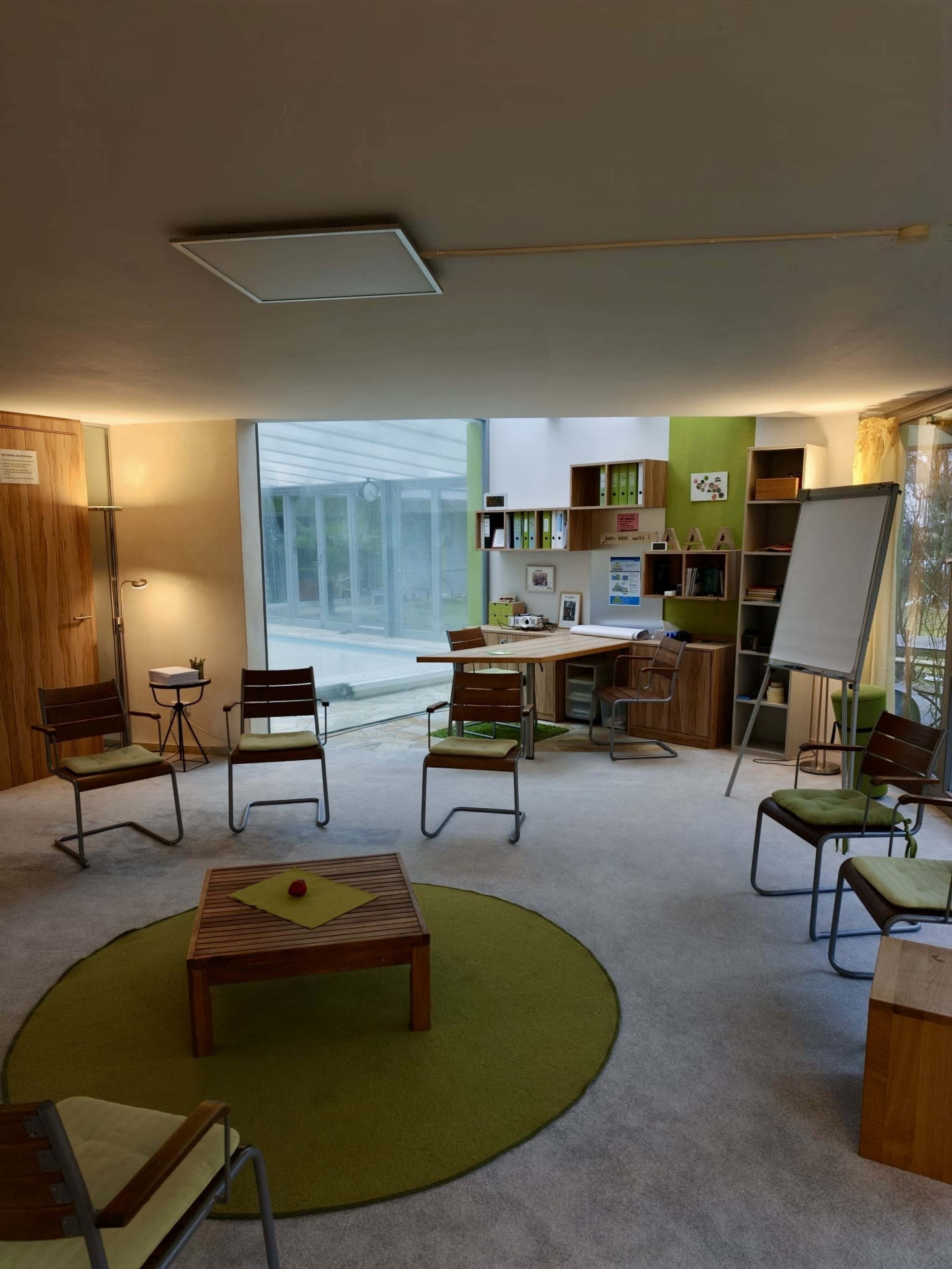 The image shows a modern office space with a round green rug, a wooden table, and several chairs arranged around it, featuring large windows that allow natural light to enter.