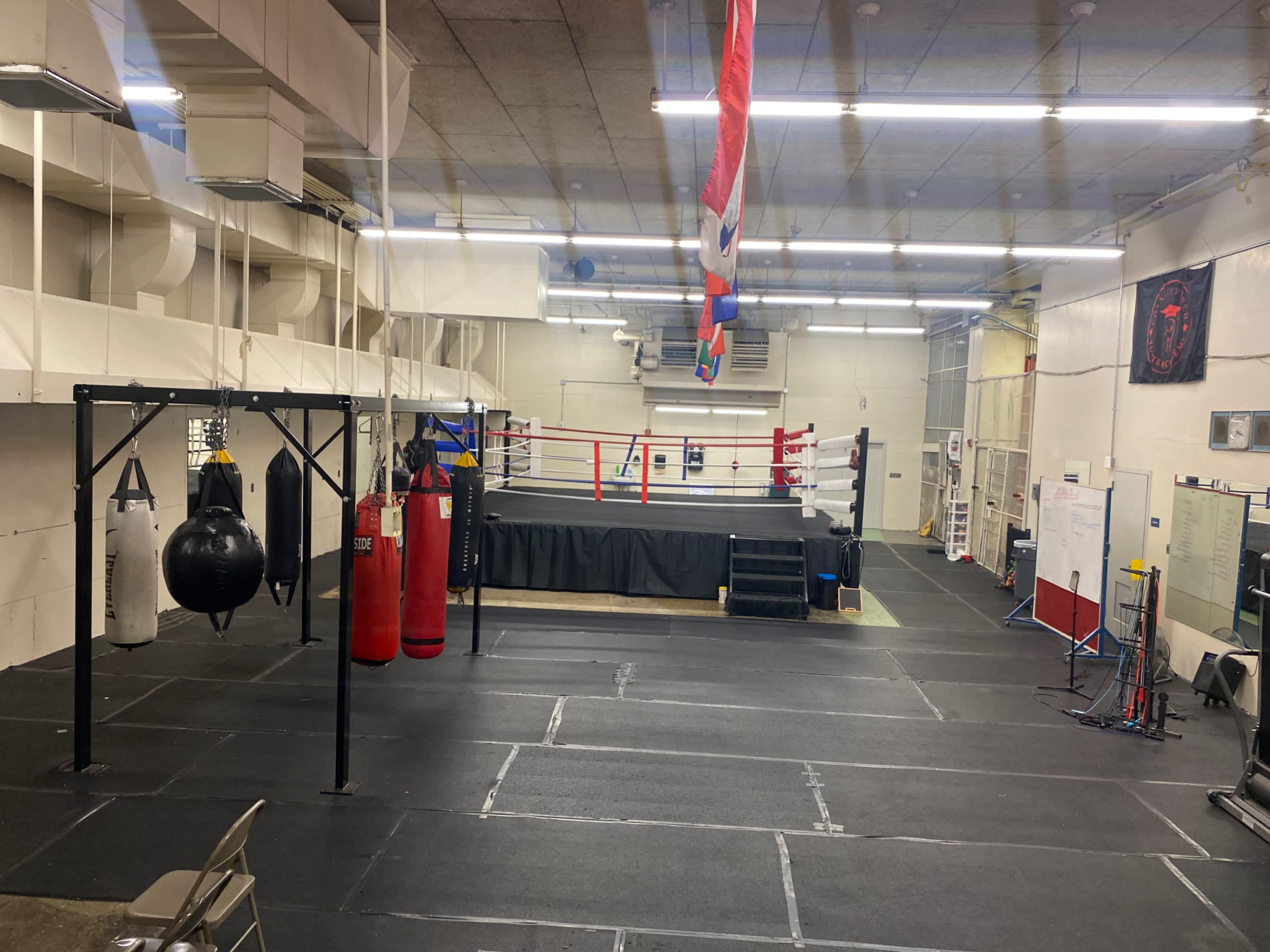 The image shows an empty boxing gym with a central ring, hanging punching bags, and mats on the floor.