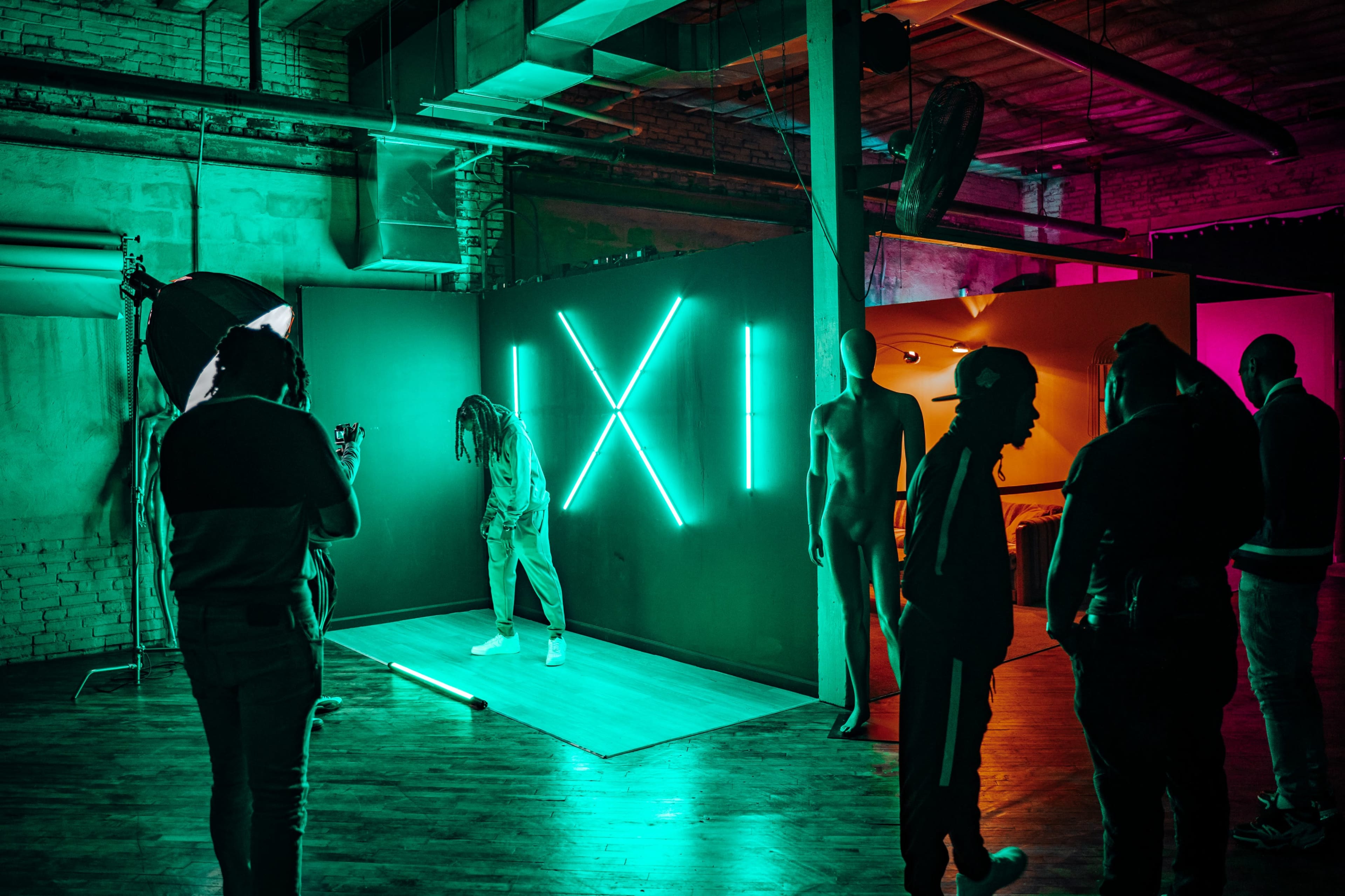 A person stands in a dimly lit studio surrounded by neon lights and silhouettes of mannequins while others observe and capture the scene.