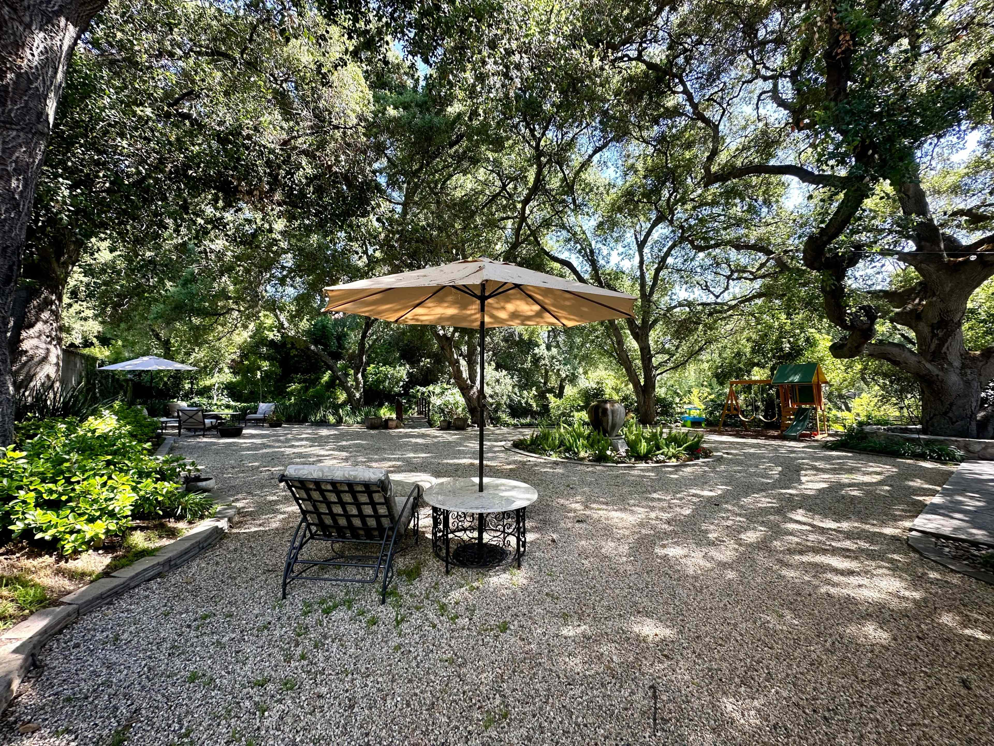 A gravel garden area features a table and chairs under an umbrella, surrounded by lush greenery and trees.