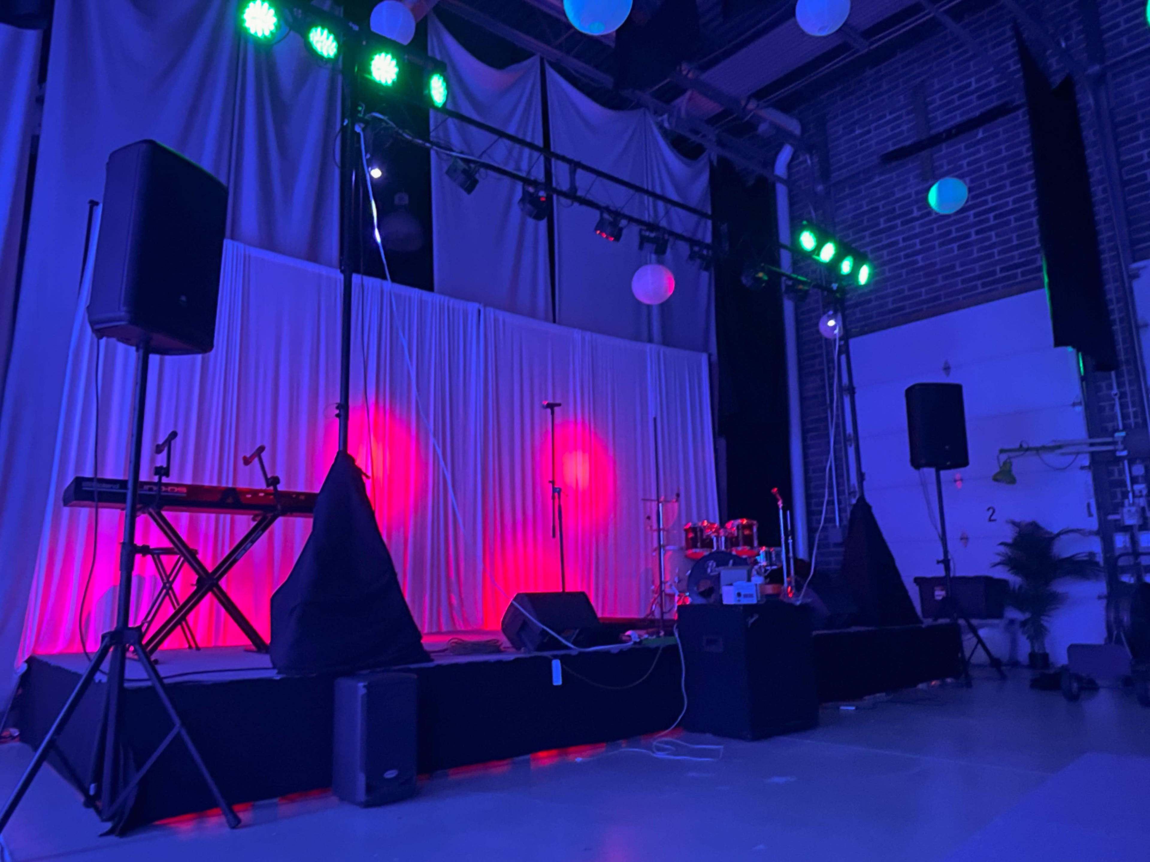 A stage is set with a keyboard, drums, and colorful lights in a dimly lit venue.