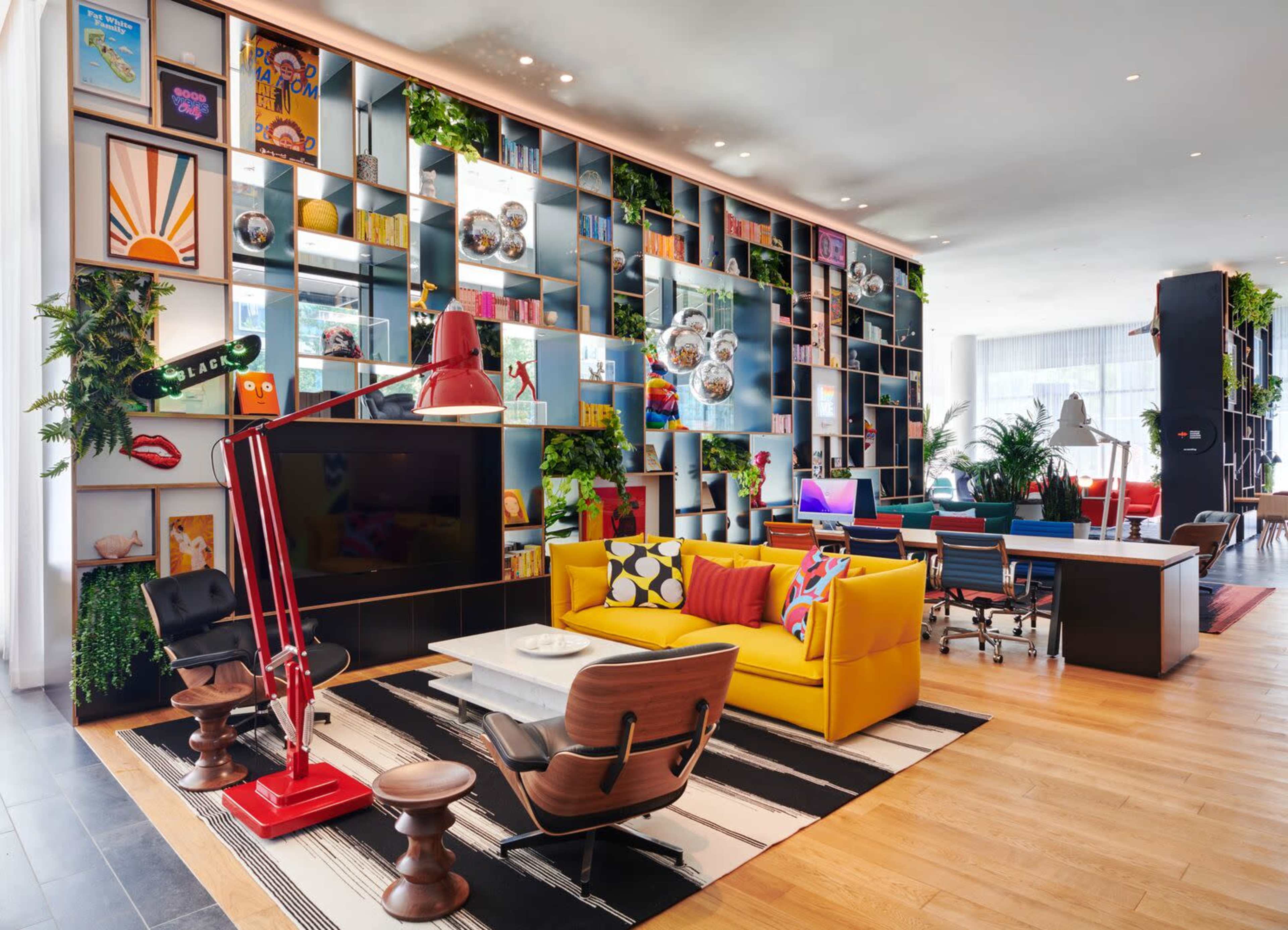 The image features a modern workspace with colorful furniture, a large bookshelf filled with decorative items, and multiple work areas.