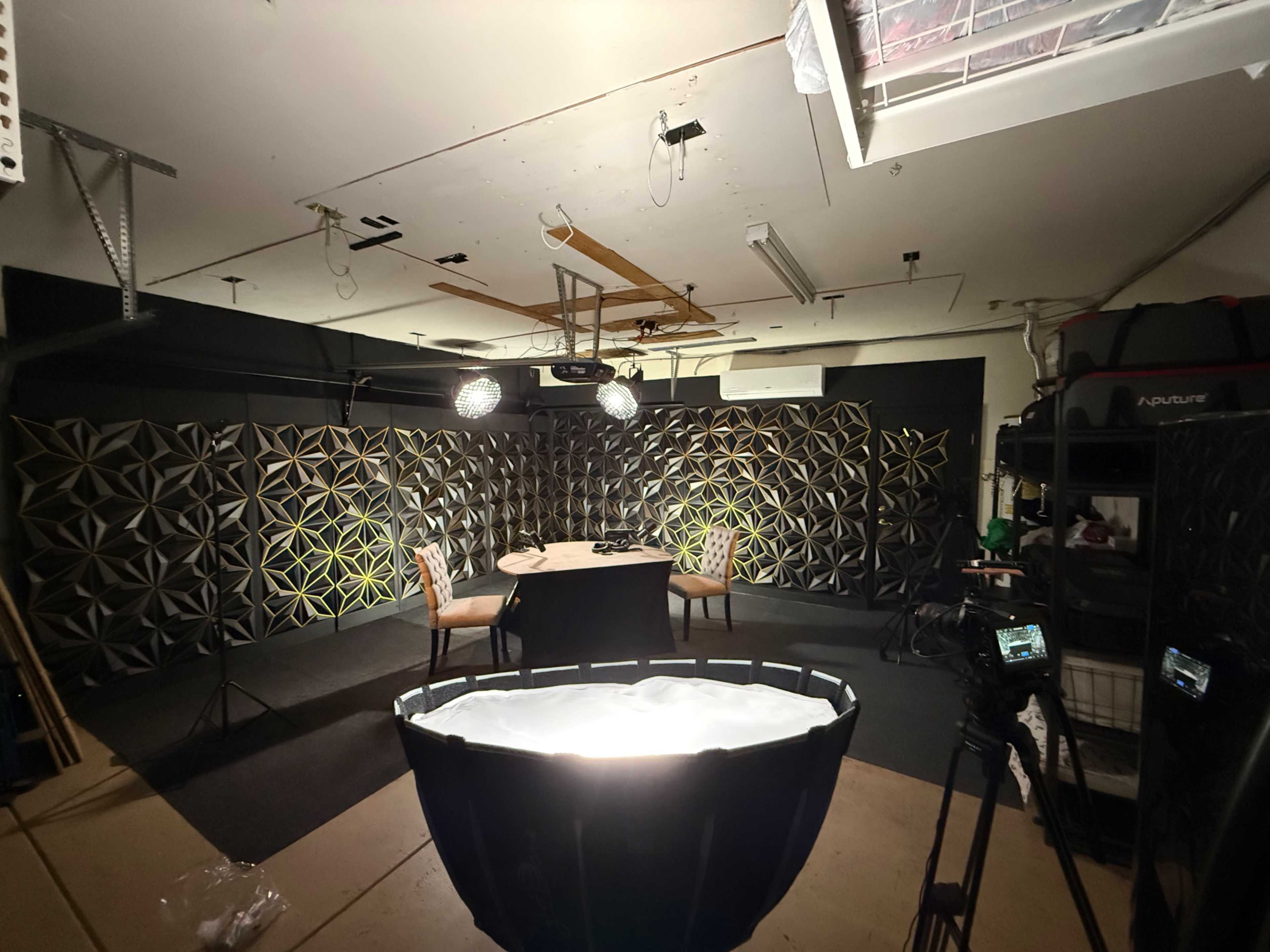 The image shows a dimly lit studio space with a round table and two chairs, surrounded by a decorative geometric wall and equipped with studio lights and cameras.