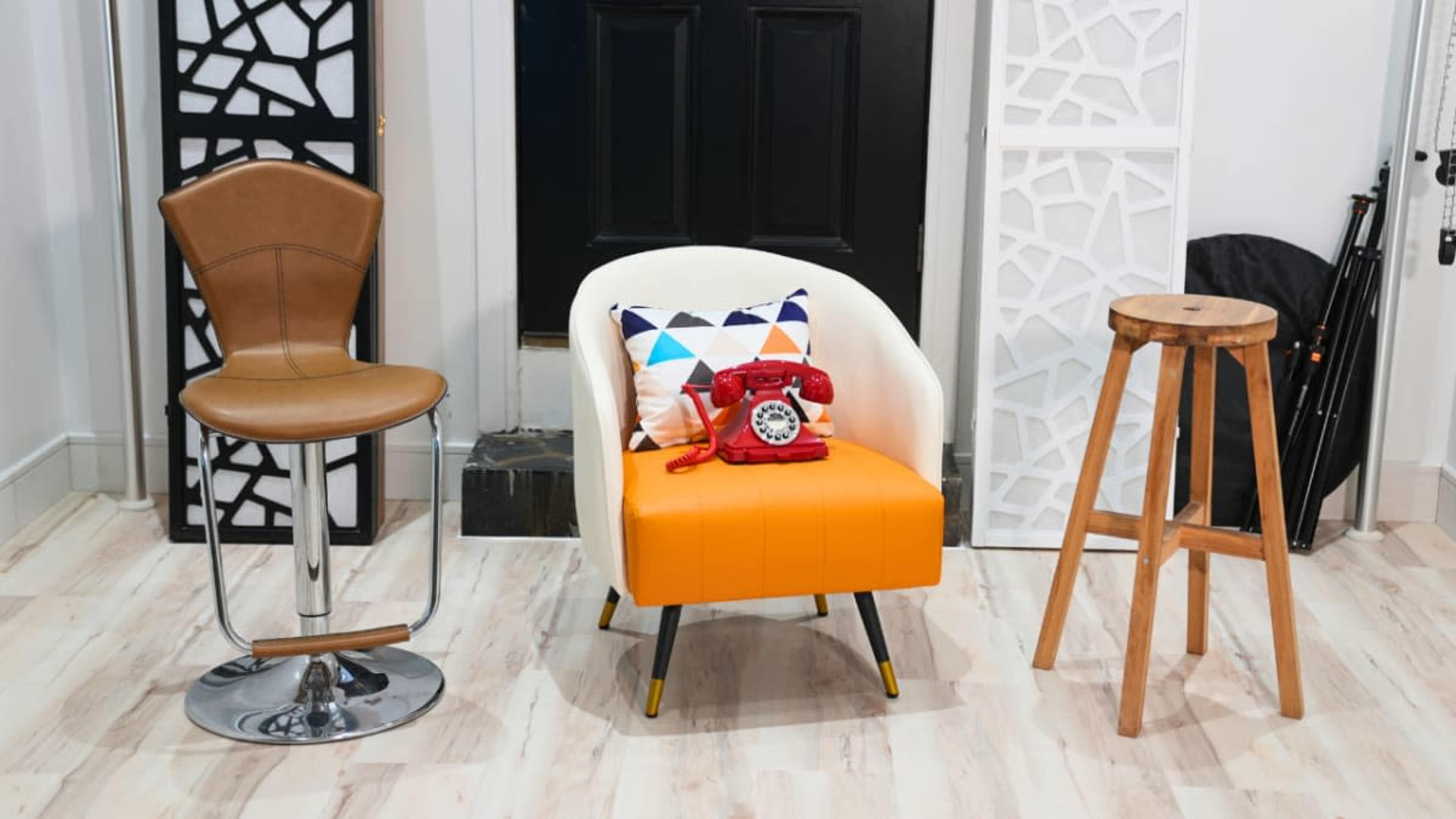 The image shows a small orange upholstered chair with a decorative pillow and a vintage telephone, flanked by a brown bar stool and a wooden stool against a black door.
