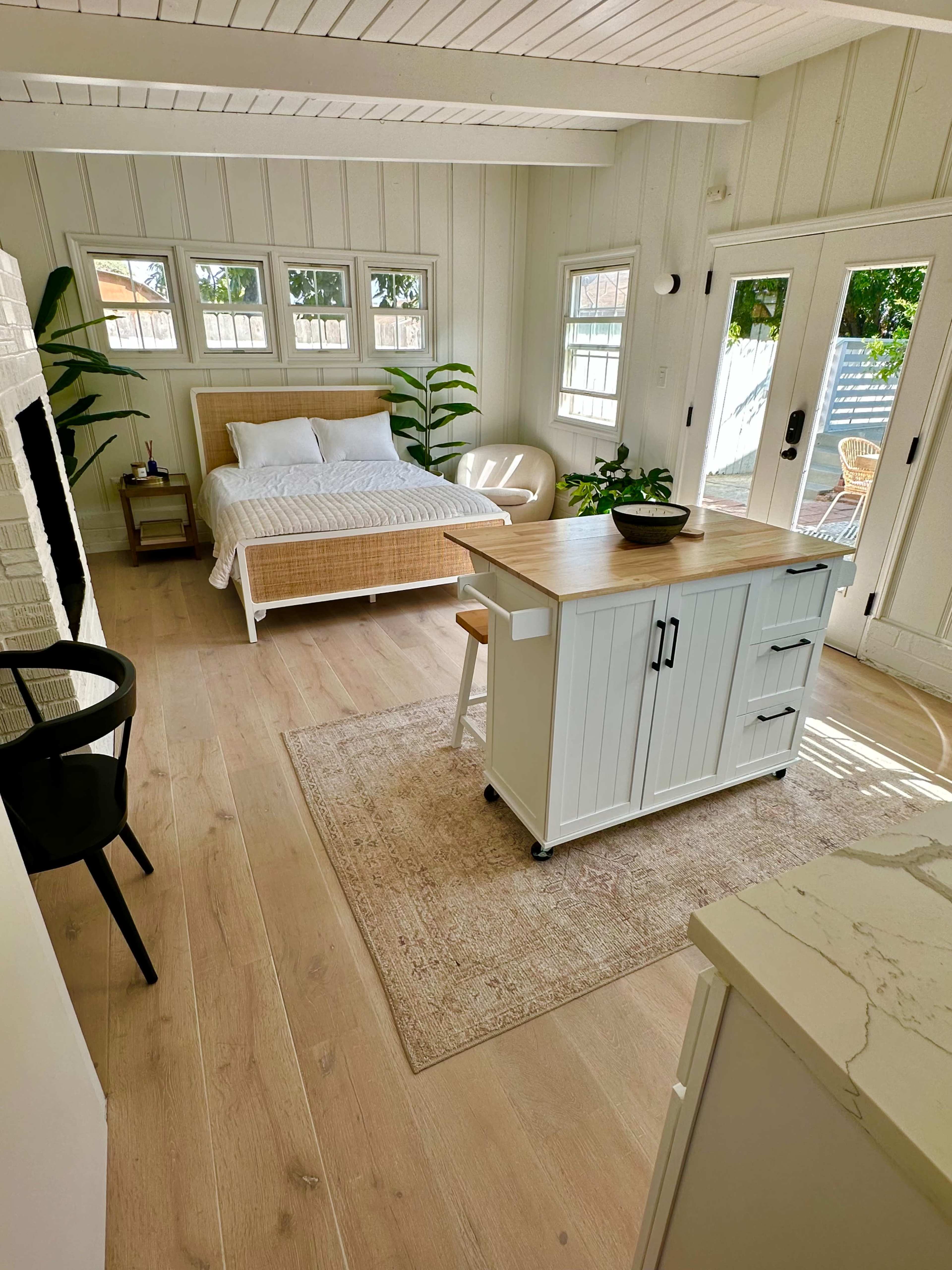 The image shows a bright, minimalist bedroom with a bed, a wooden cart, a chair, and large windows allowing natural light.
