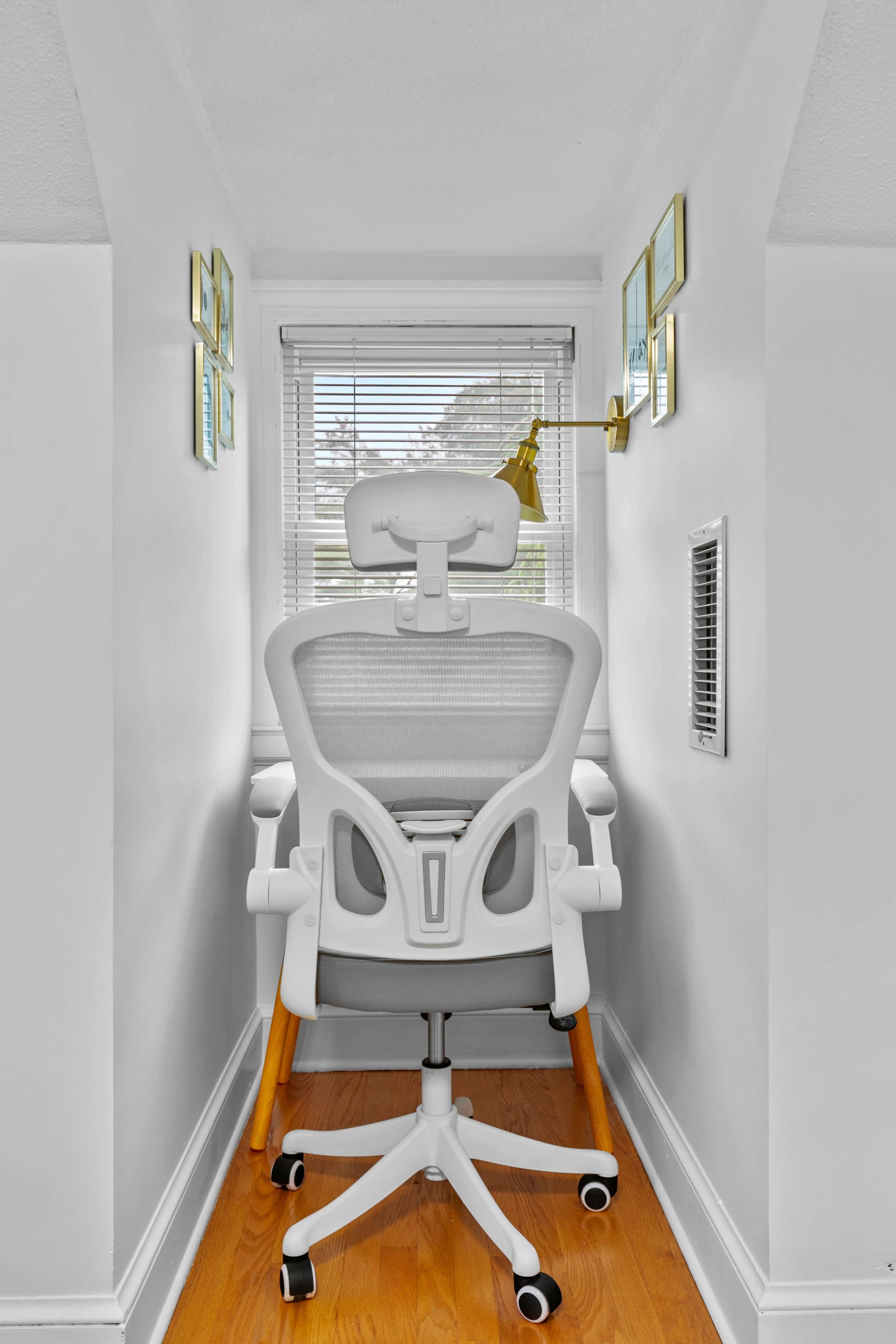 A white ergonomic office chair is positioned against a wall, facing a window with blinds, in a narrow, well-lit space.