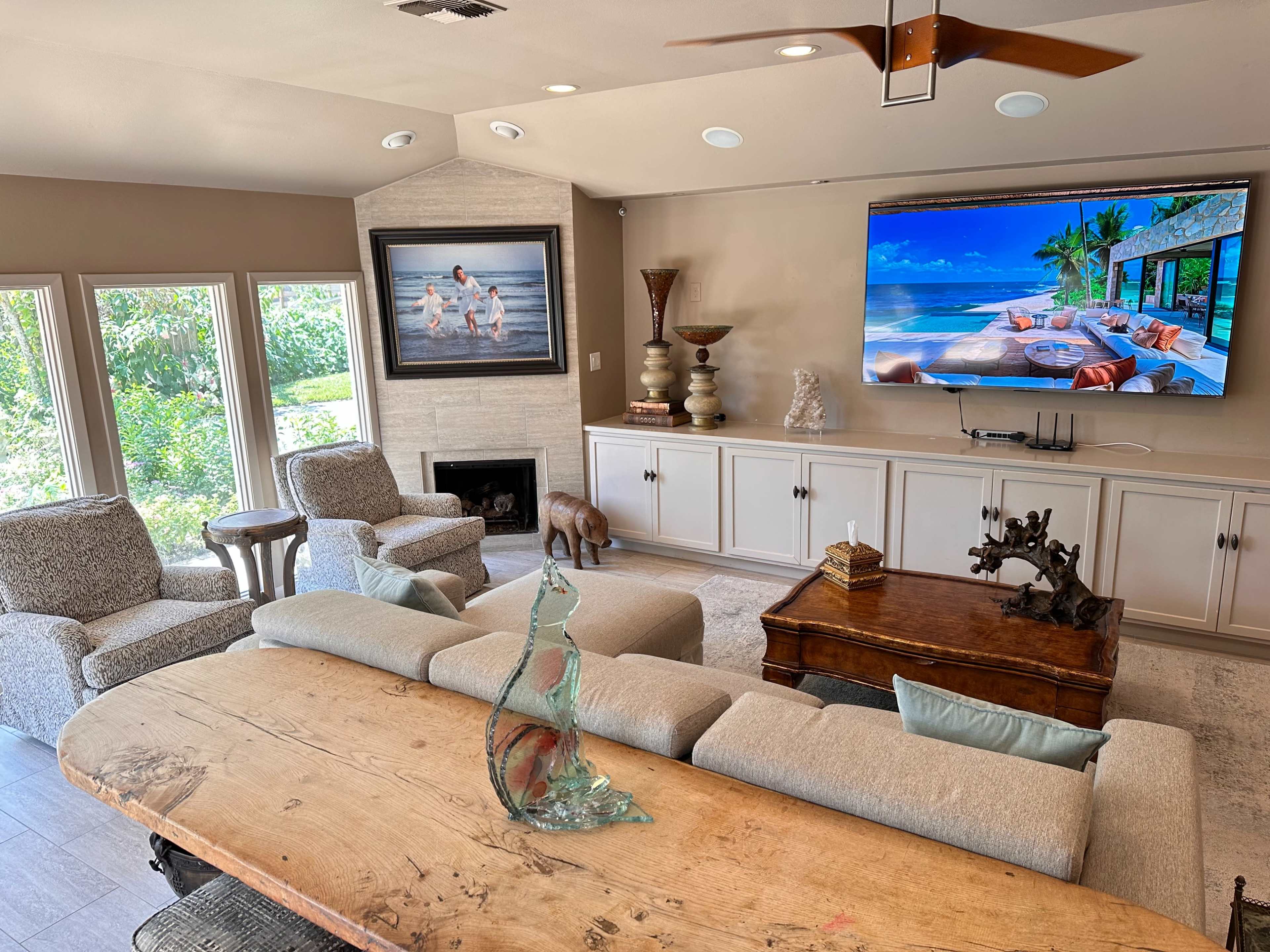 A spacious living room features a large television mounted on a light-colored wall, a wooden coffee table, and comfortable seating arranged around a fireplace, with natural light streaming in from multiple windows.