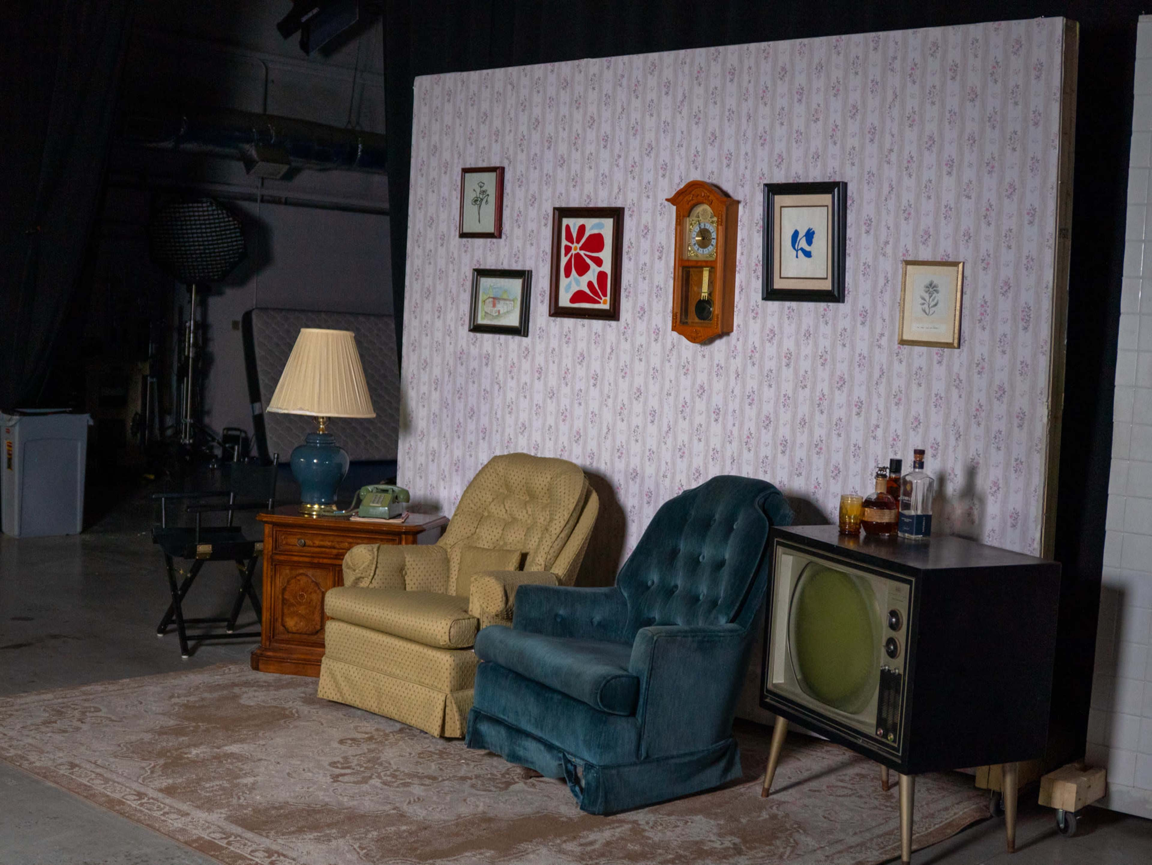 The image shows a room setup with two armchairs, a vintage television, a side table with a lamp, and a wall adorned with framed art and a clock.