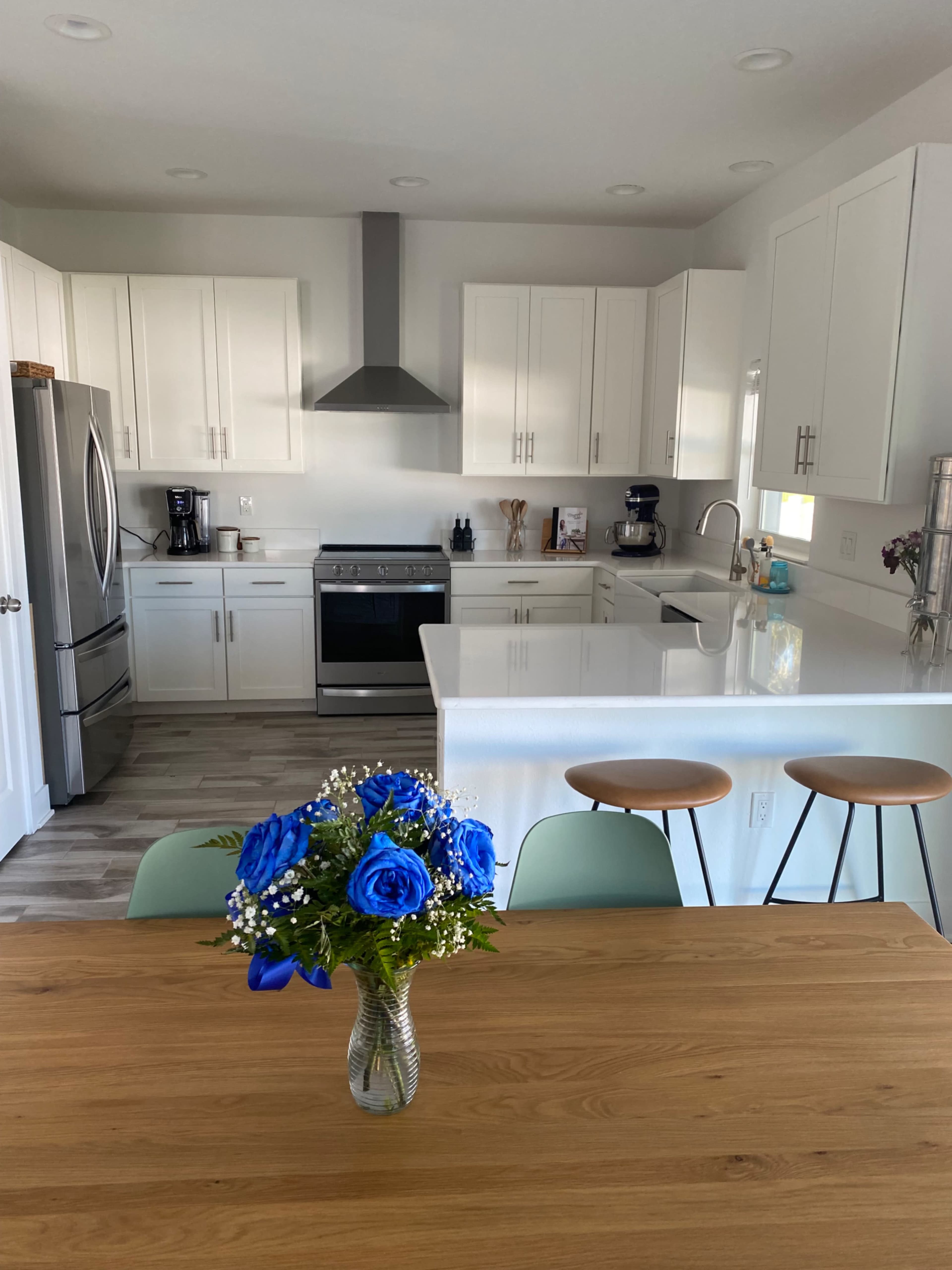 A modern kitchen features white cabinetry, stainless steel appliances, a large island with bar stools, and a dining table with a vase of blue roses.