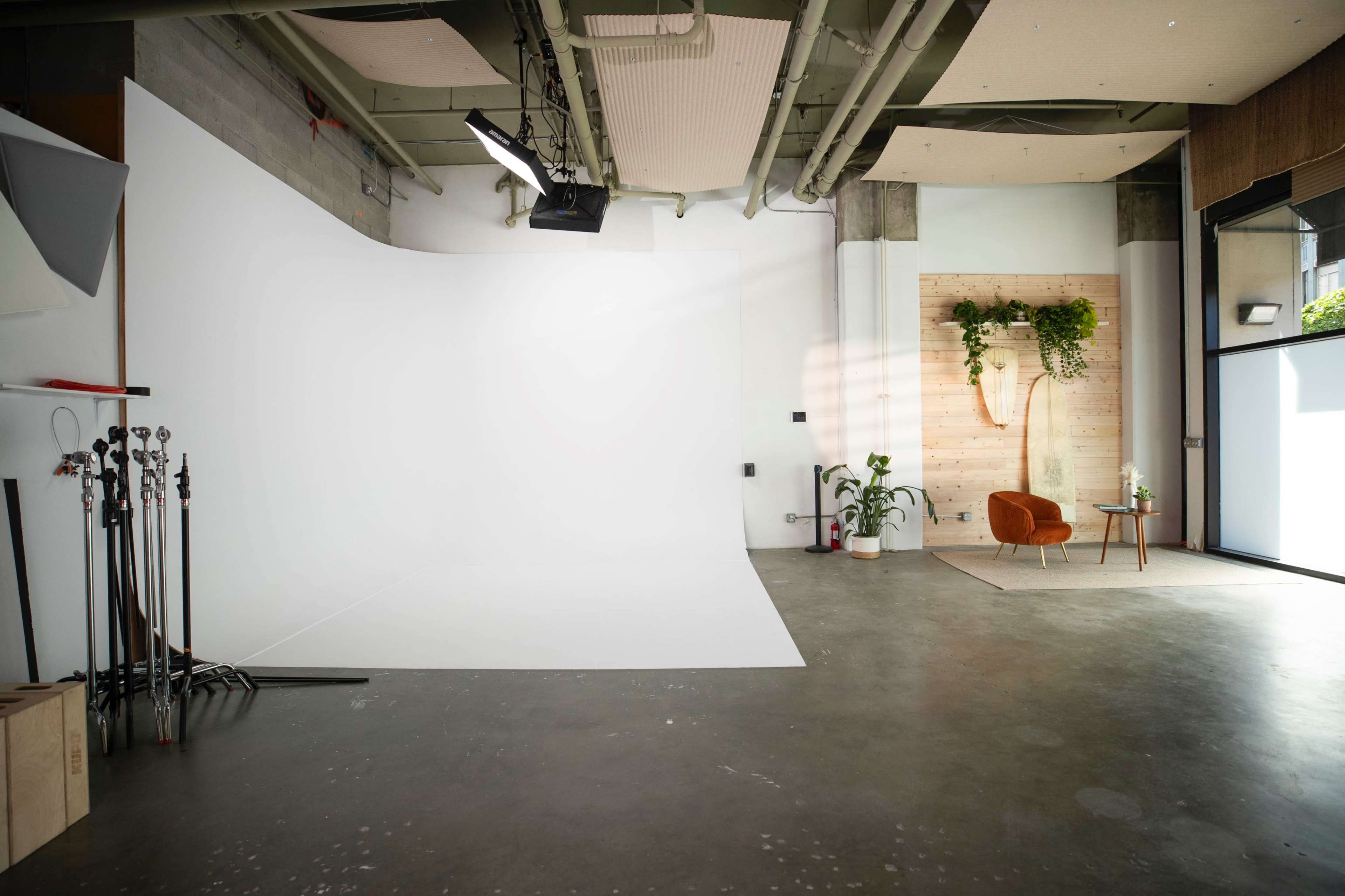 A minimalist studio space with a large white backdrop, concrete flooring, and a cozy orange chair beside a small table and greenery on the wall.