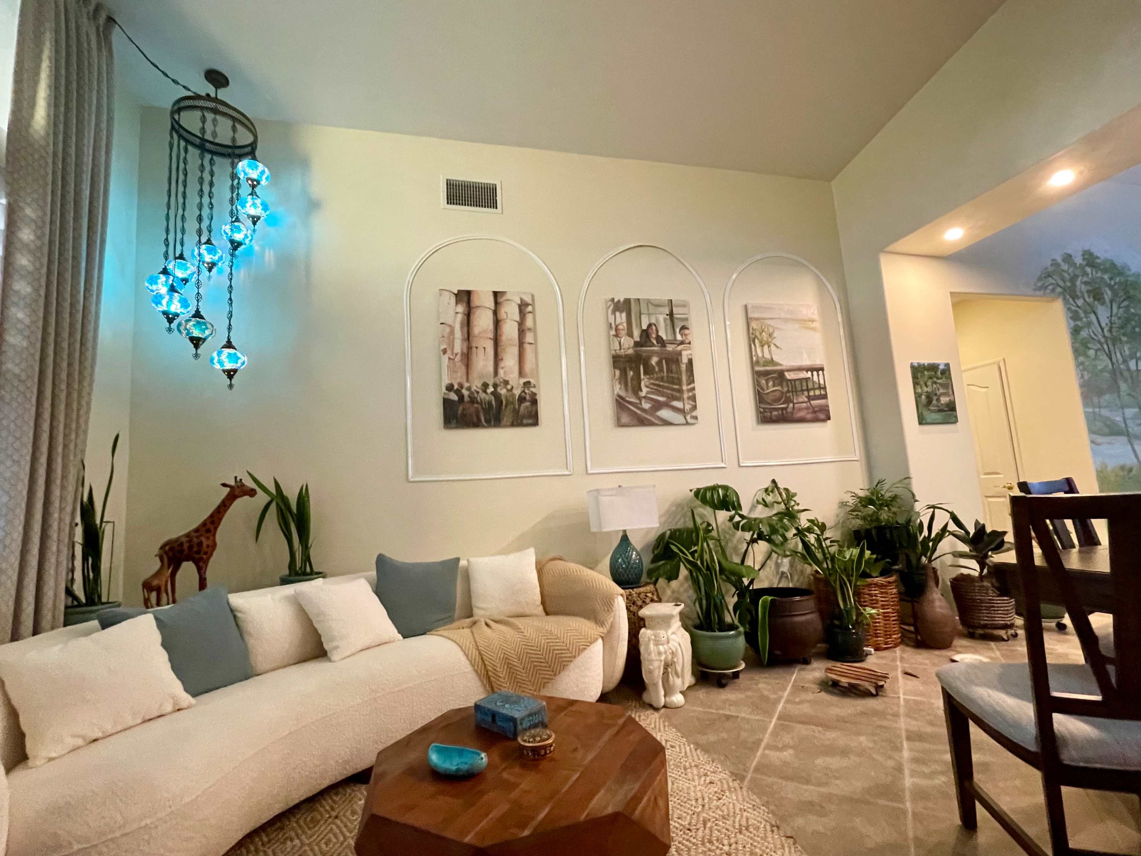 The image shows a cozy living room with a curved sofa, decorative plants, and framed artwork on the wall, illuminated by a unique chandelier.