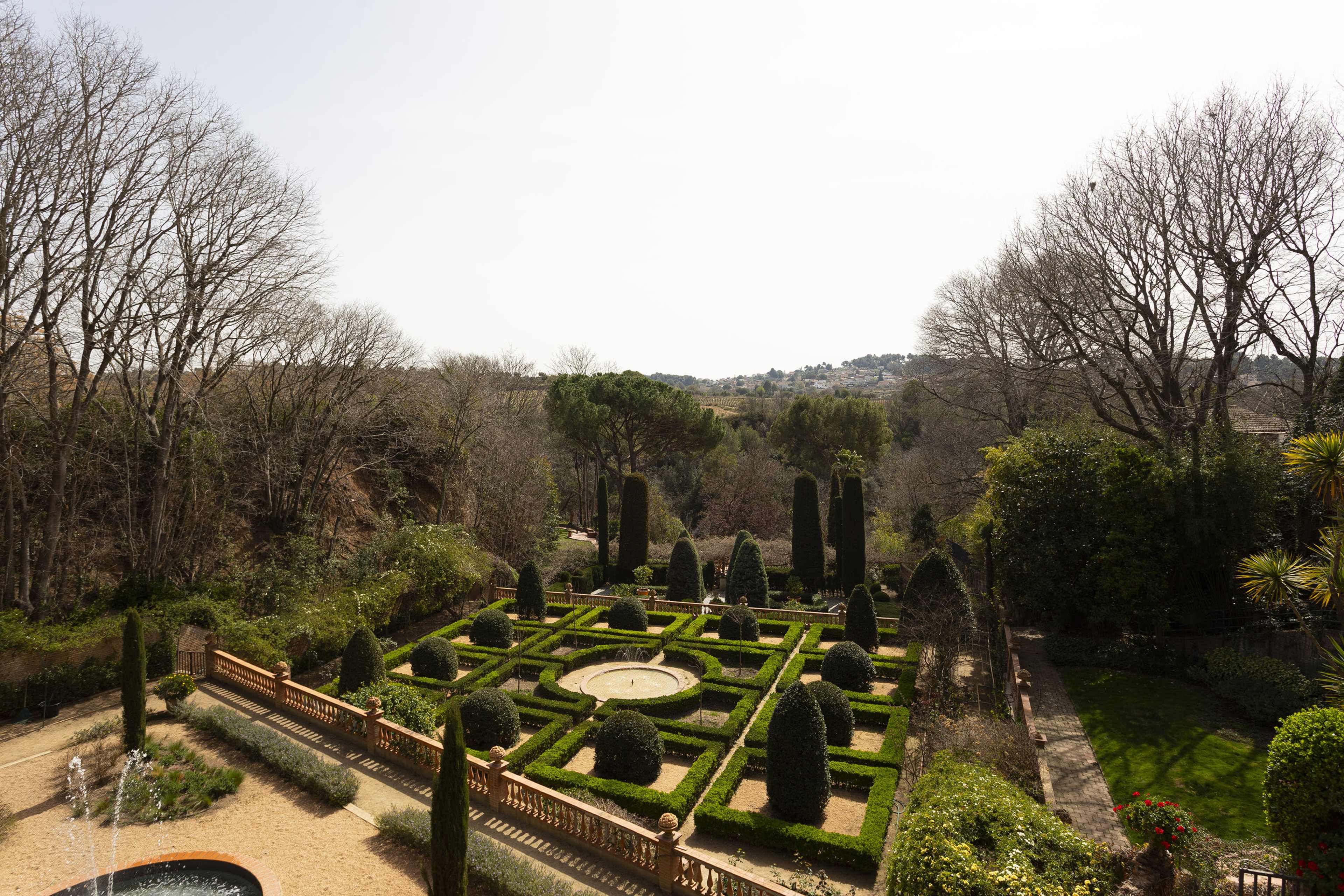 A well-manicured garden features neatly arranged hedges, topiary, and geometric flowerbeds, surrounded by bare trees and distant hills.