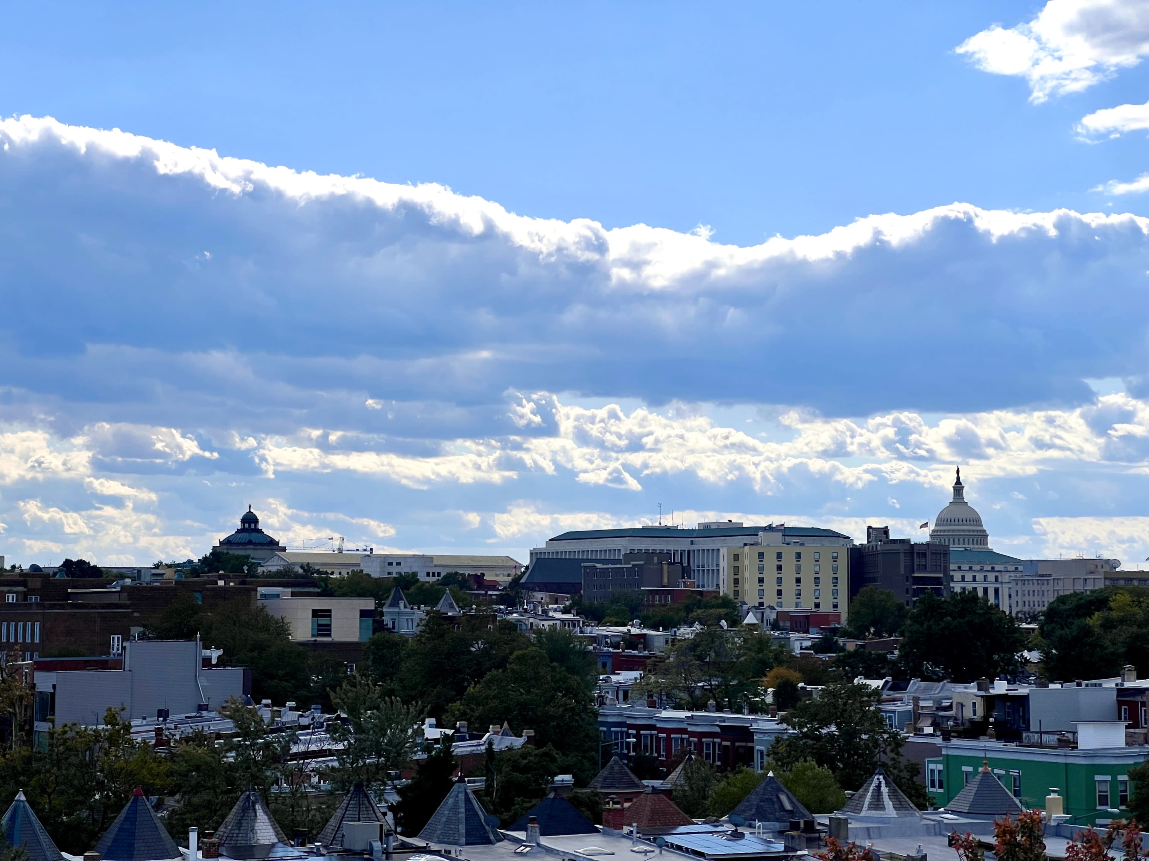A panoramic view of a city skyline featuring distinct buildings, including a domed capitol and a large complex, under a partly cloudy sky.