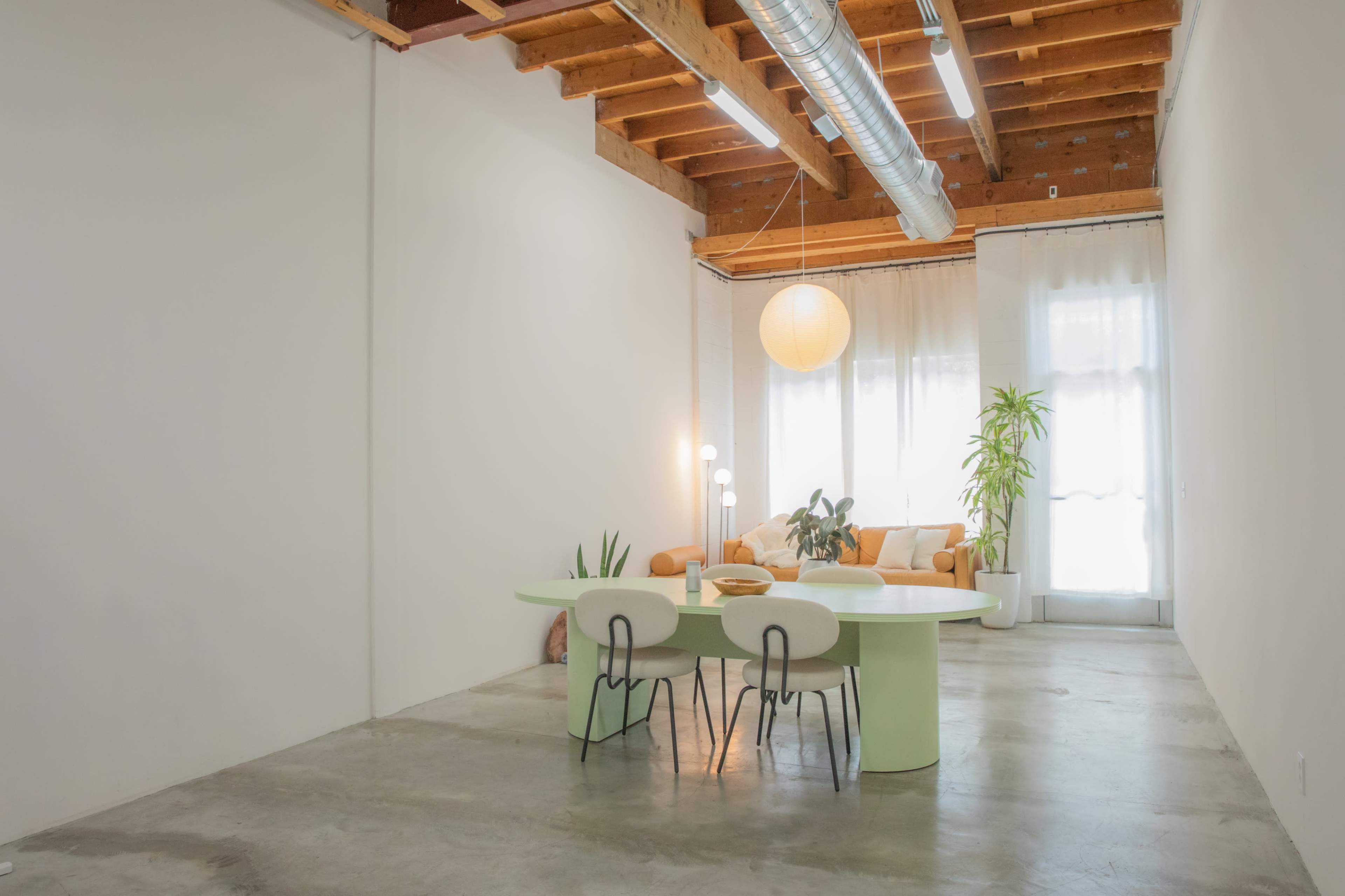 A modern, minimalist interior with a green dining table surrounded by black and white chairs, a sofa in the background, and large windows allowing natural light.