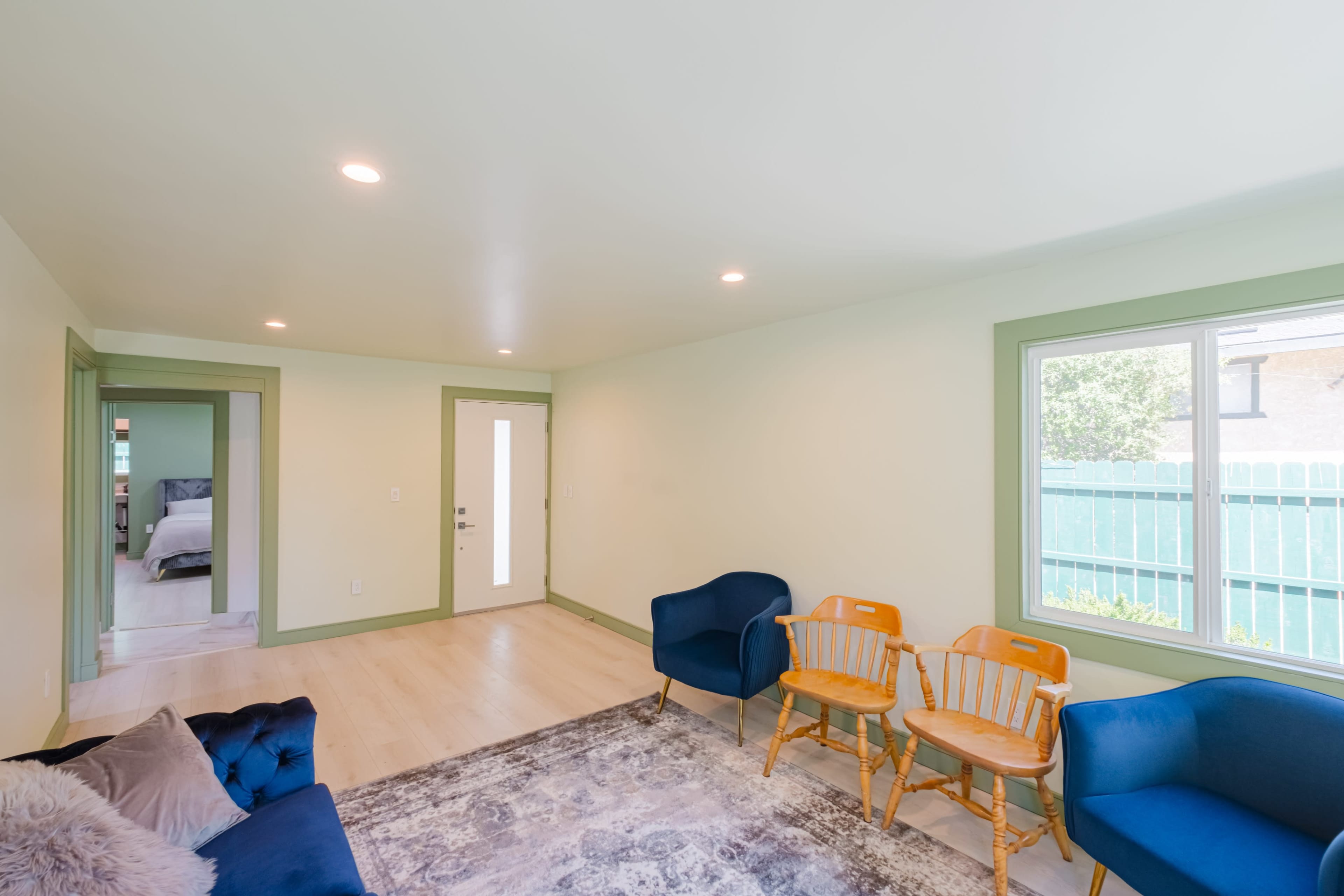 A bright, minimalist living room features two blue chairs, two wooden armchairs, and a door leading to another room.