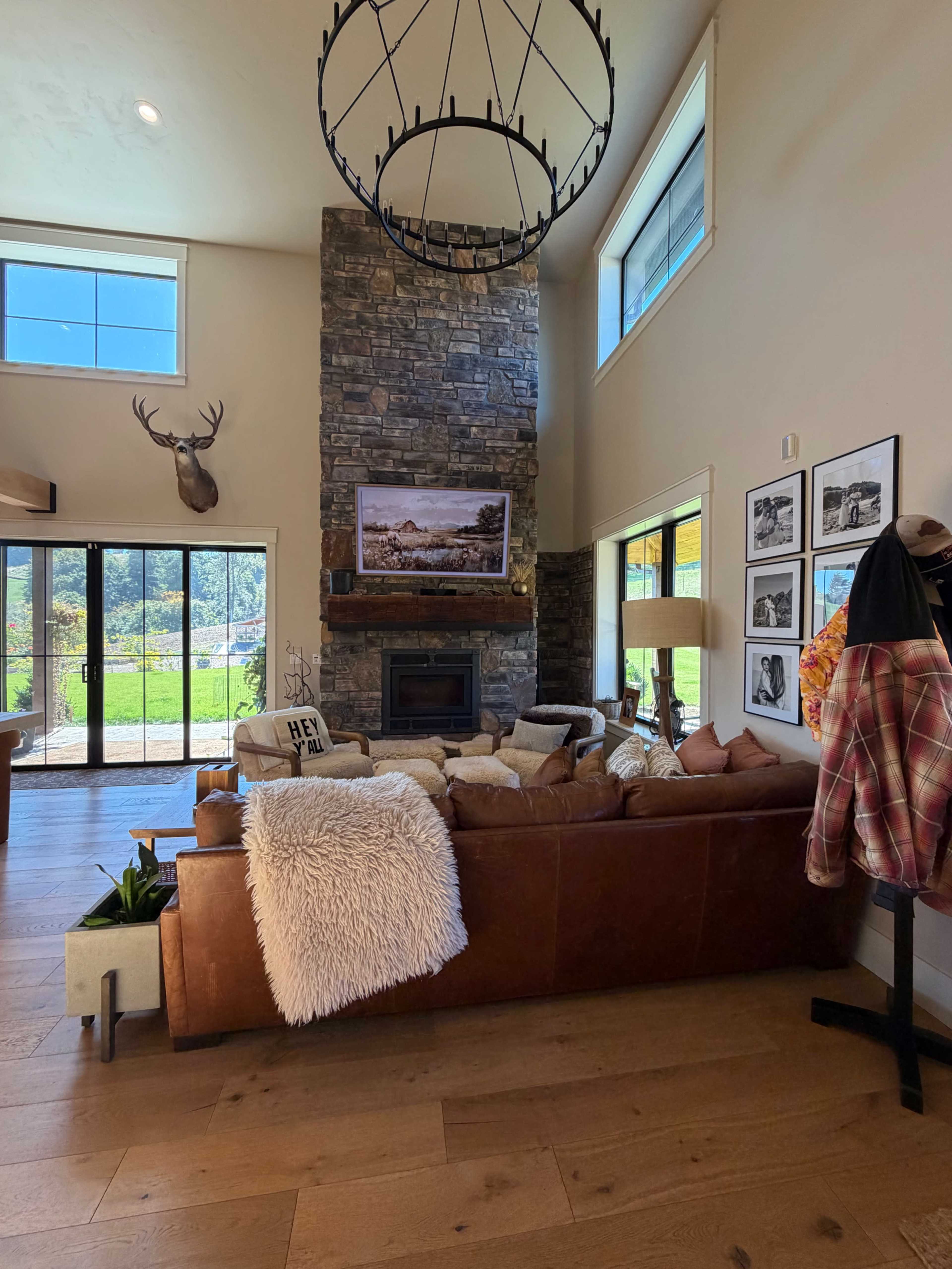 The image shows a modern living room featuring a large stone fireplace, a leather sofa adorned with pillows, and a coffee table with a small plant, all set against a backdrop of large windows that offer a view of greenery outside.