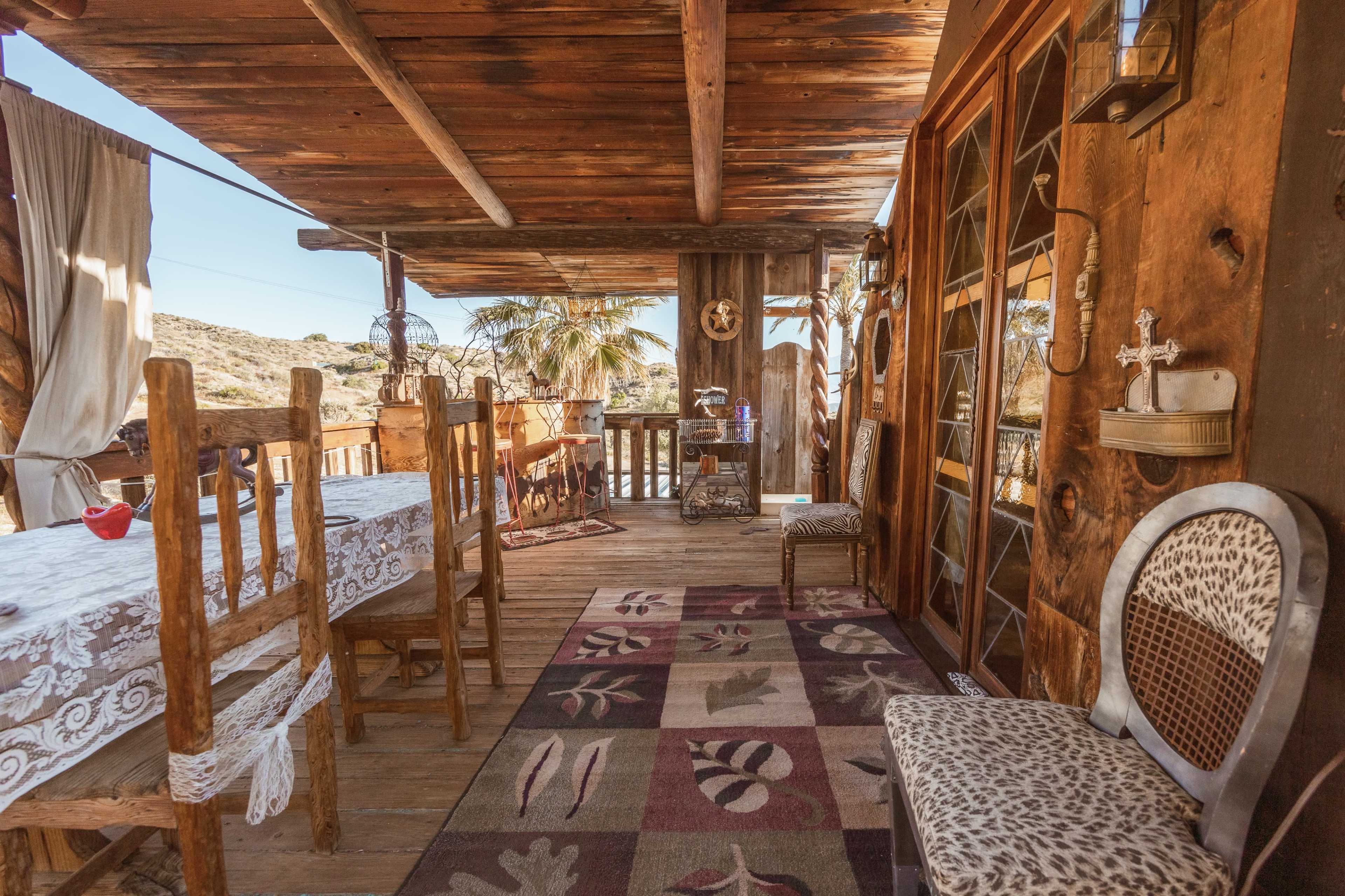 The image shows a rustic wooden porch featuring a table with chairs, a patterned rug, and various decorative elements, overlooking a scenic outdoor view.