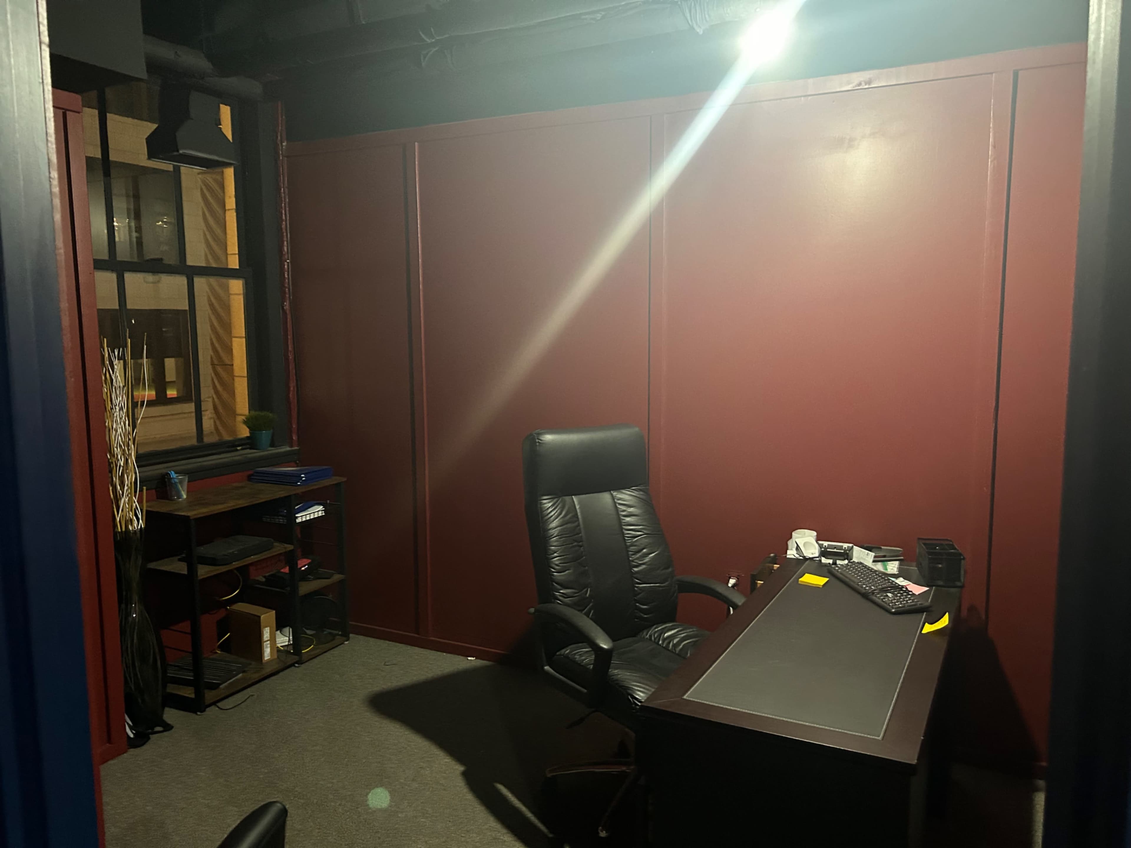 A dimly lit office space features a black leather executive chair and a dark wooden desk against maroon walls, with a shelf and window visible in the background.