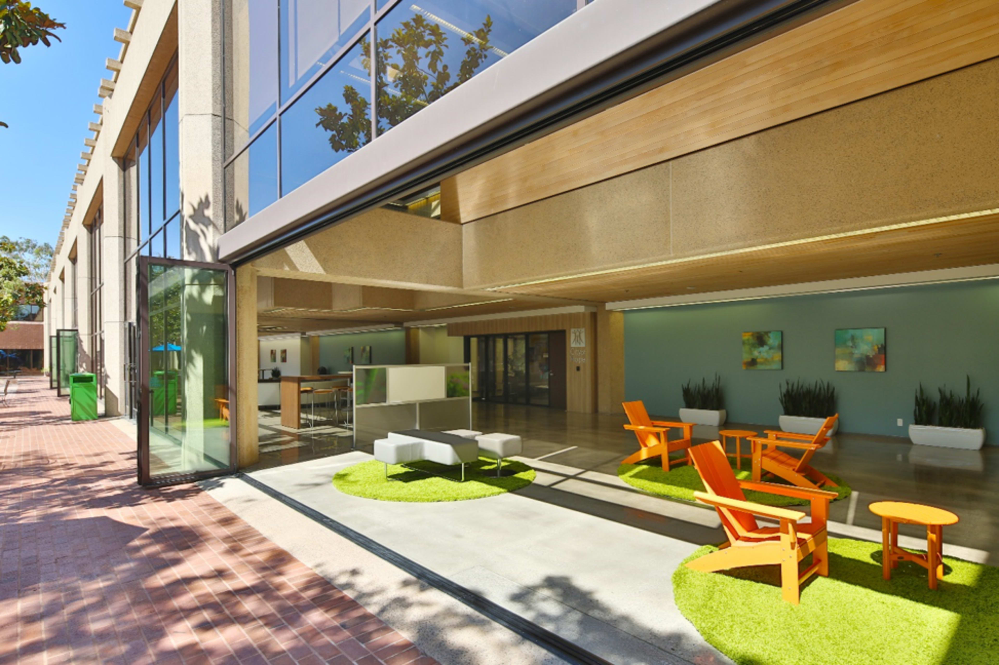 A modern building features large glass walls opening to a patio with orange chairs and green grass-like matting.
