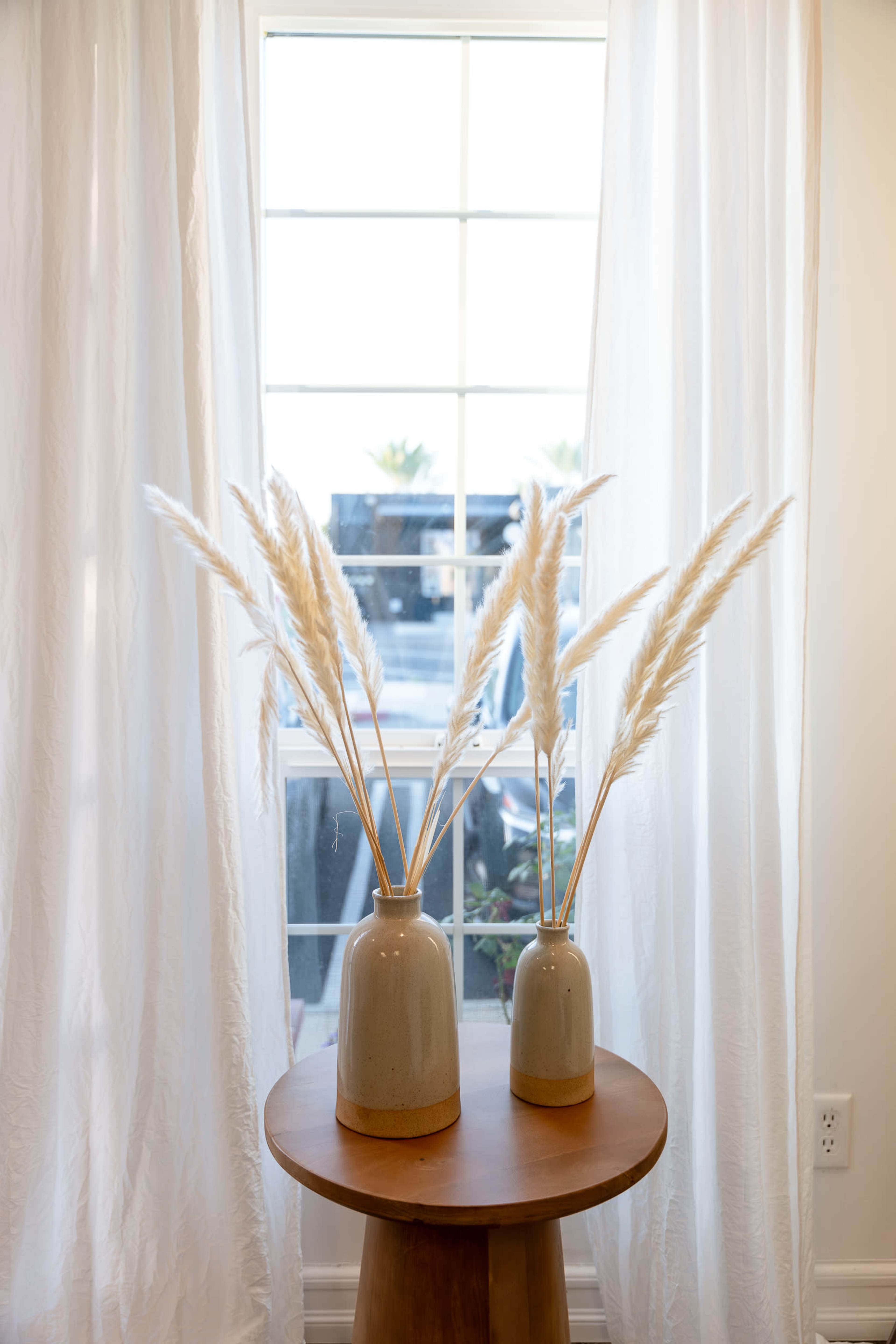 Two ceramic vases with pampas grass stand on a wooden table next to a large window draped with white curtains.